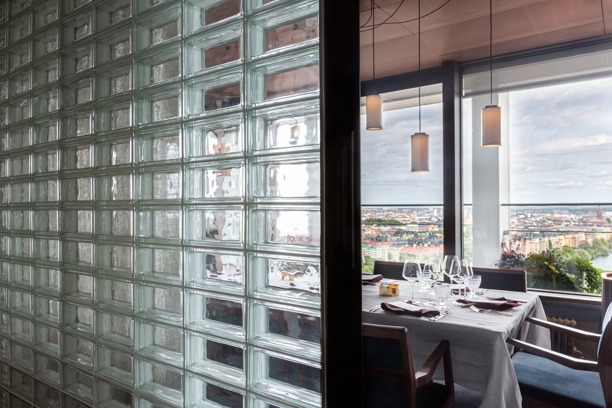 Ett modernt konferensrum eller restaurang med glasväggar och stora fönster som visar en stadsvy med byggnader och ett vattendrag. Inomhus finns ett dukat bord med glas, bestick och servetter, samt några hängande lampor i projektet La Tour i Stockholm
