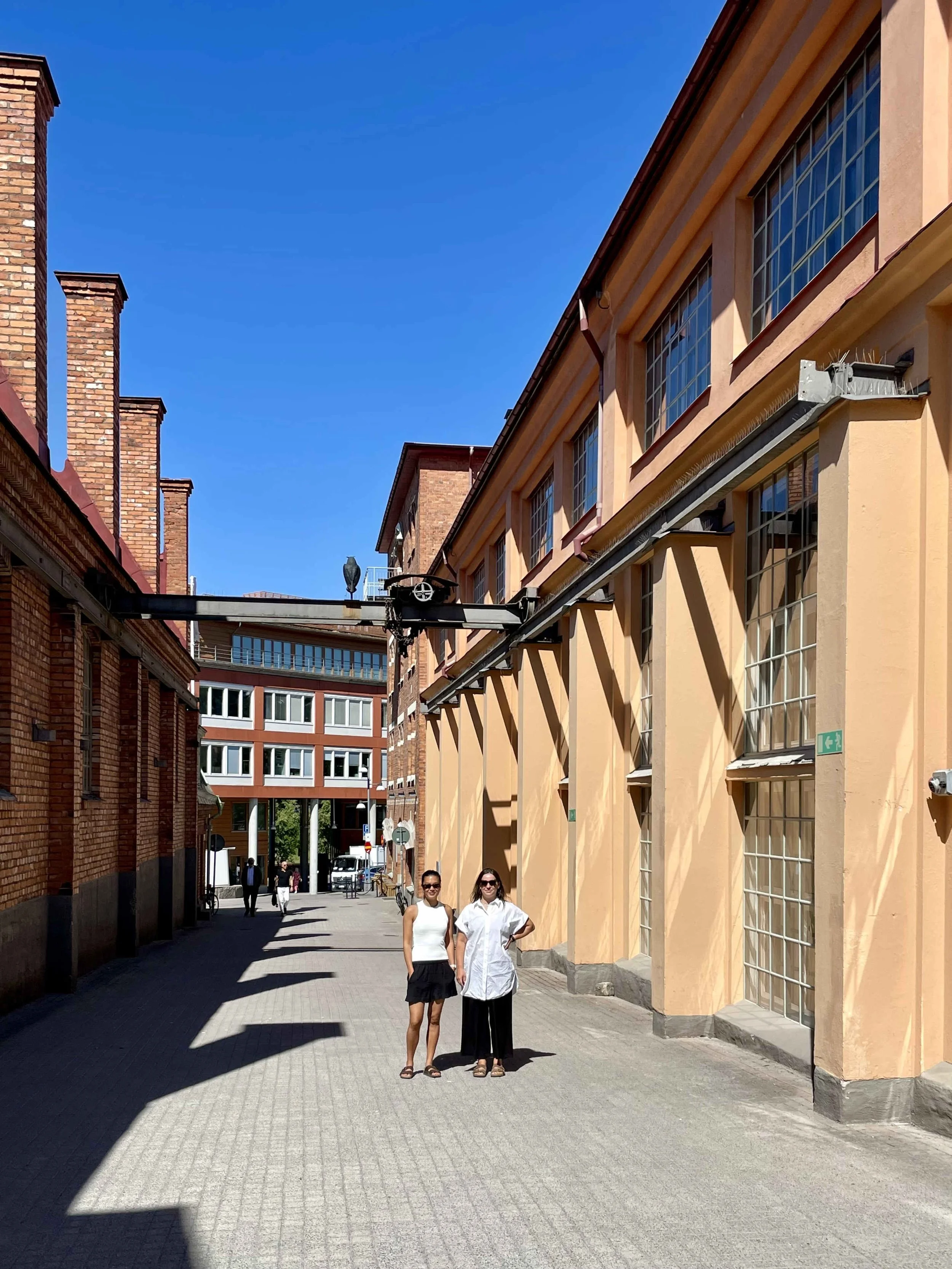 Två kvinnor står på en stadsgata, en i vitt linne och svart kjol, den andra i vit skjorta och svarta byxor, under en klar blå himmel omgivna av äldre byggnader med stora fönster.