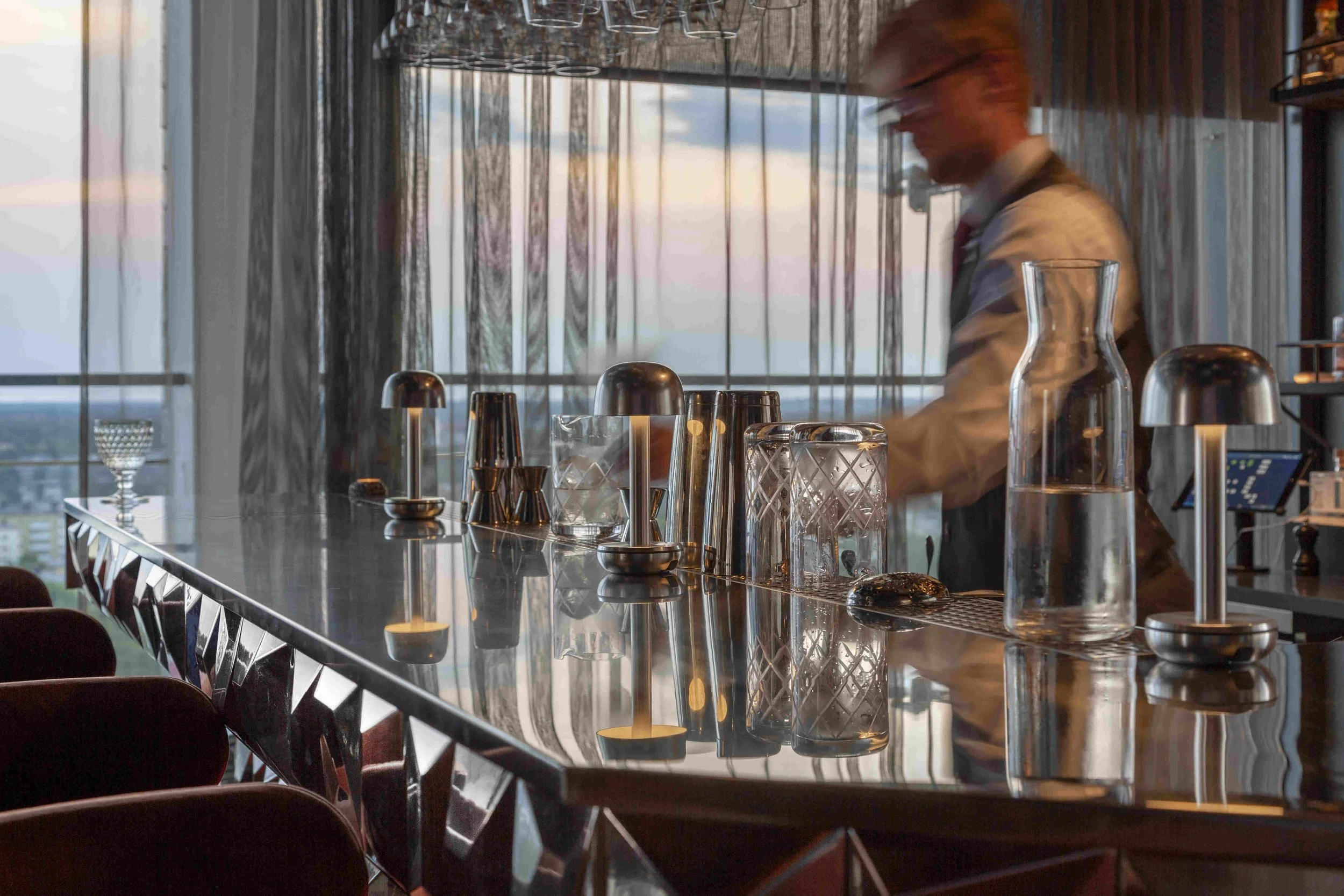 En bartender står bakom en svart bardisk med glas och bartillbehör, med stora fönster och en stadssilhuett i bakgrunden i projektet Le Bar i Stockholm