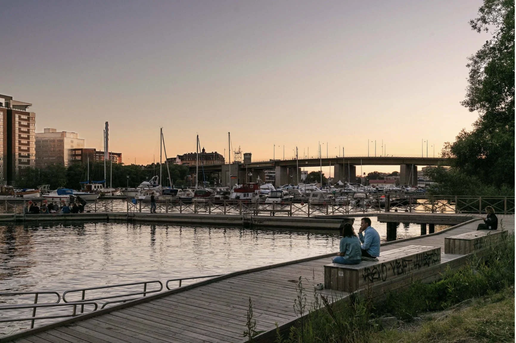 Många båtar och segelbåtar i en liten hamn vid solnedgången, med en bro och stadsmiljö i bakgrunden, samt några personer som sitter och pratar vid vattnet.