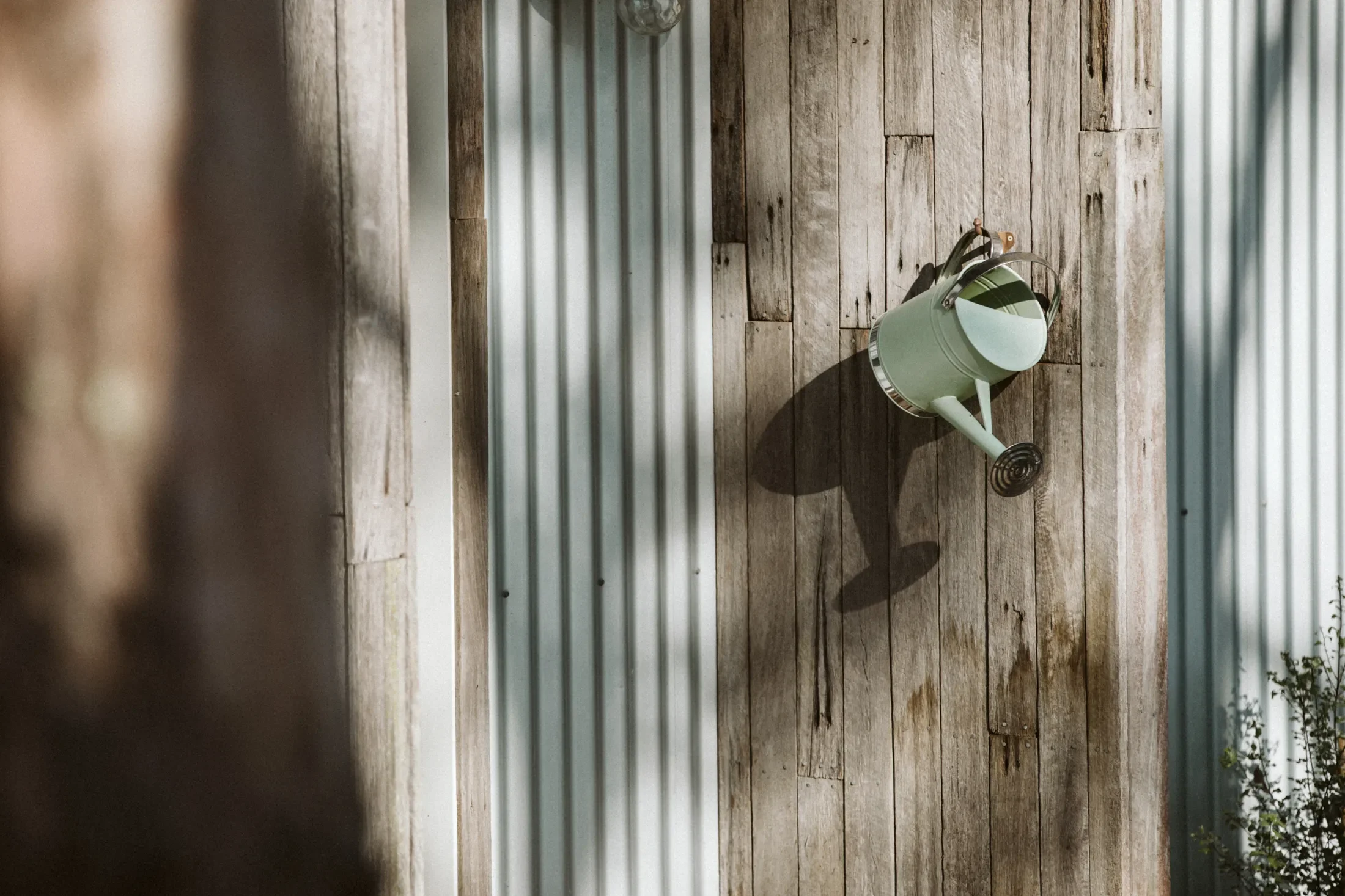A light green watering can hanging on a weathered wooden wall next to a metal siding wall.