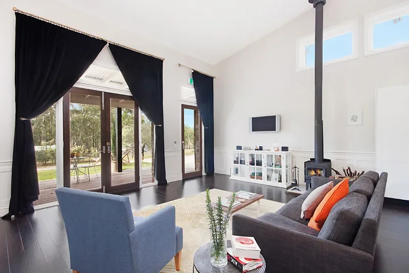 Living room with dark hardwood floor, light-colored walls, large glass doors with dark curtains, a gray couch with orange and white pillows, a blue armchair, a white media console, a wood stove, and a small coffee table with books and a plant.
