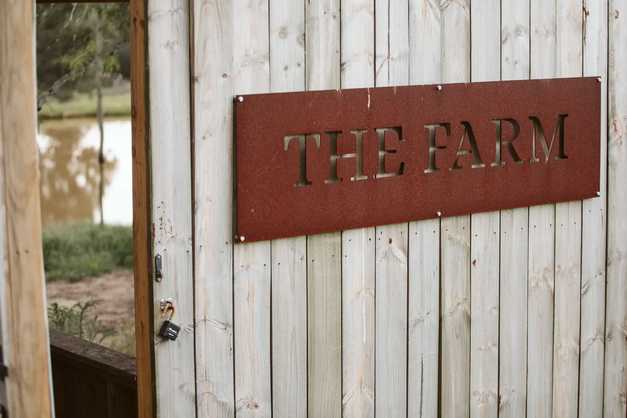 A wooden fence with a red metal sign that reads 'THE FARM' is shown in the image. The fence has vertical planks, and there are trees and a body of water visible in the background.