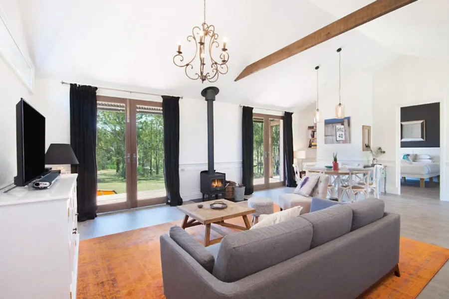 Living room with a gray sofa, a coffee table, a wood-burning stove, and a large window with black curtains showing greenery outside.