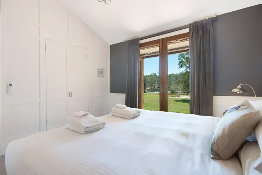 A bedroom with white walls and a window showing trees and blue sky outside, neatly made bed with white linens, pillows, and folded towels, gray curtains, and a small wall-mounted lamp.