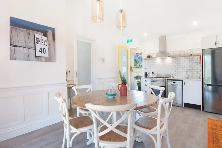 Bright kitchen and dining area with a round wooden table surrounded by white chairs, a red teapot, and a glass dish in the center, white cabinets, a stainless steel refrigerator, and a white subway tile backsplash.
