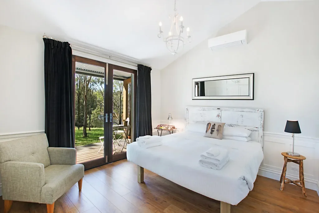A bright bedroom with a large bed, white bedding, and a decorative pillow, with a small wooden side table and black lamp, a beige armchair, and glass doors leading to a wooden patio with outdoor chairs and a view of trees.