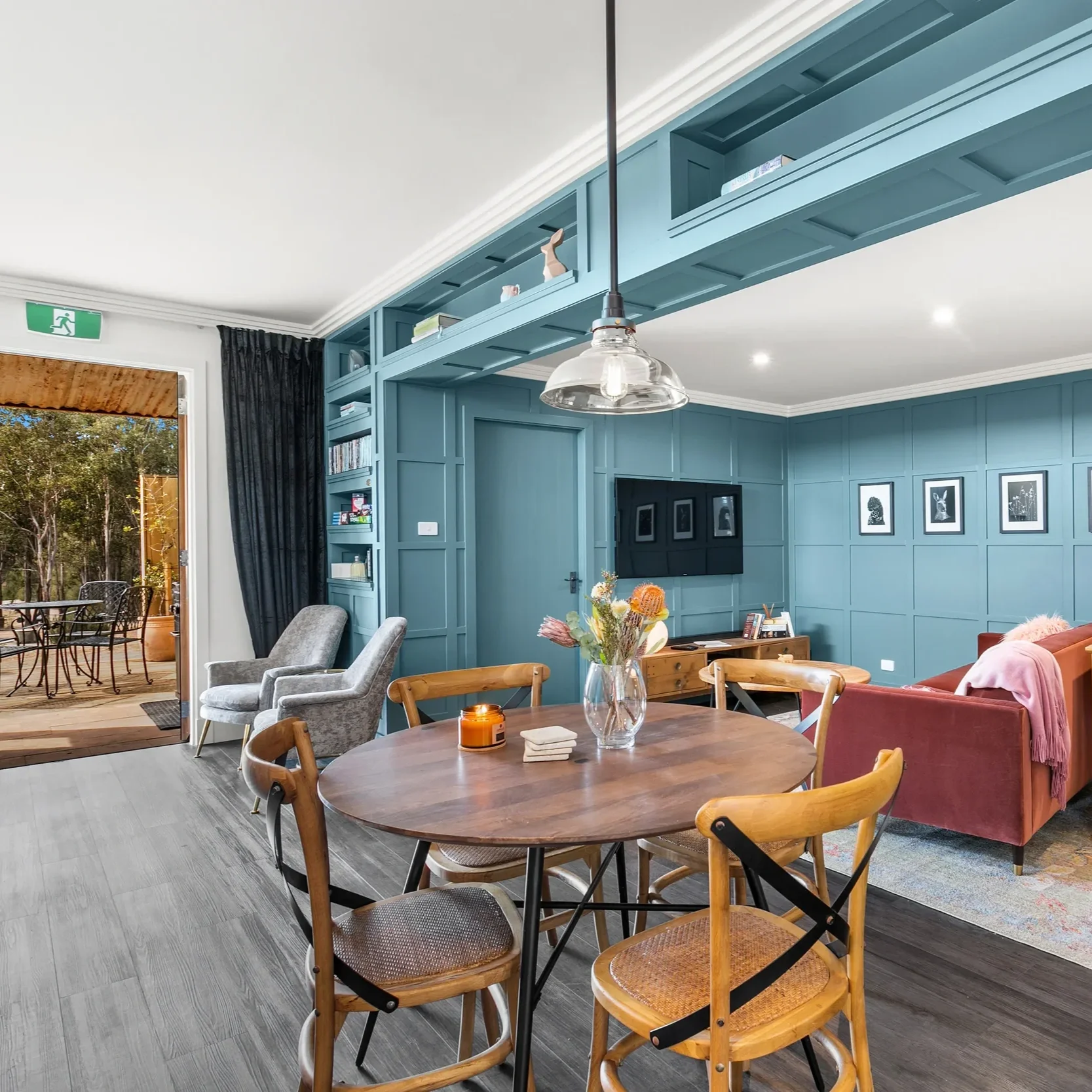 The image shows a modern living room and dining area with blue accent walls, a wooden dining table with chairs, a pink sofa, a TV on the wall, a glass pendant light, and a sliding glass door leading to a patio with outdoor furniture and trees.