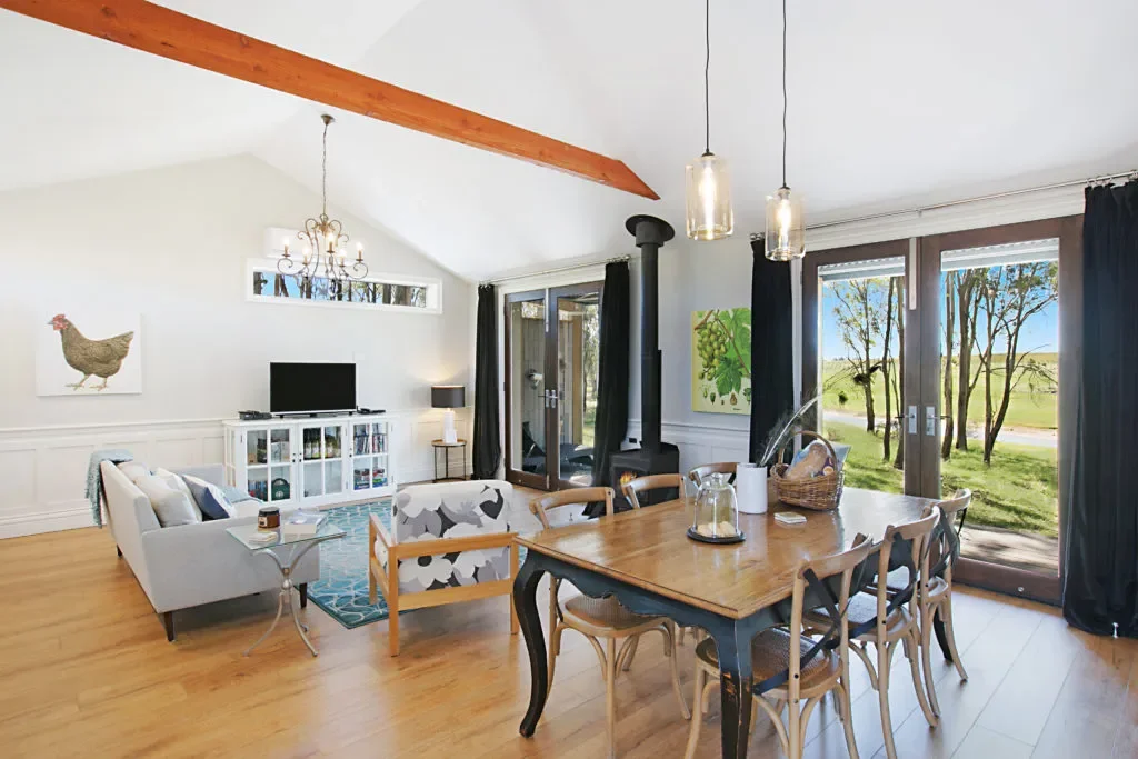 Living room with wooden dining table, black curtains, white walls, and large glass sliding doors showing outdoor trees and lawn