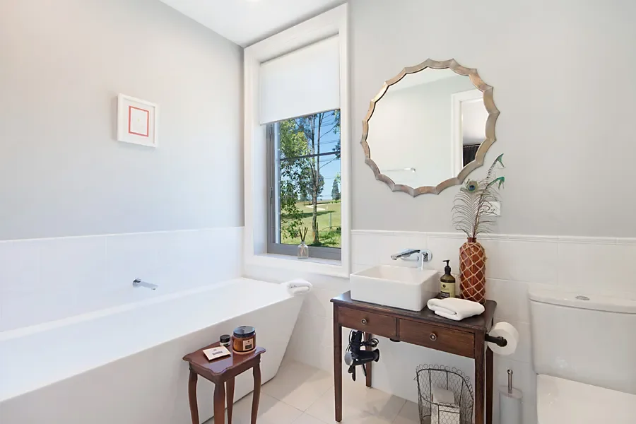 Modern bathroom with a bathtub, a window showing greenery outside, a wooden side table, a small sink with accessories, a decorative mirror, and a toilet.