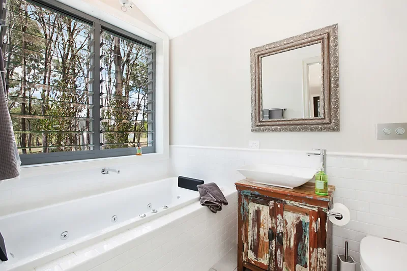 Bright bathroom with a large window, a white bathtub with jets, a rustic wooden cabinet with a vessel sink, a mirror above the sink, and a bottle of soap.