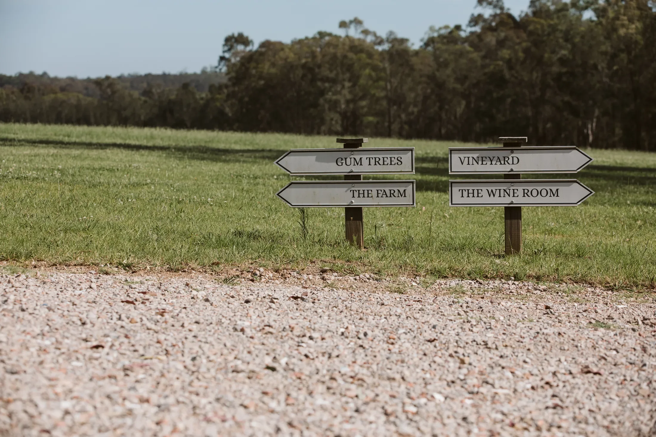 Signpost on a grassy field pointing to different locations: Gum Trees and The Farm on the left, Vineyard and The Wine Room on the right, with trees in the background.