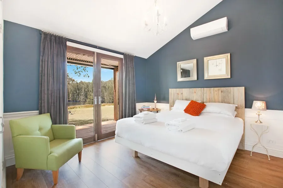 A cozy bedroom with a large bed, white bedding, and an orange pillow, against a dark blue wall with two framed pictures. There's a green armchair near sliding glass doors that open to a natural outdoor view. Small bedside lamps are on each side of th