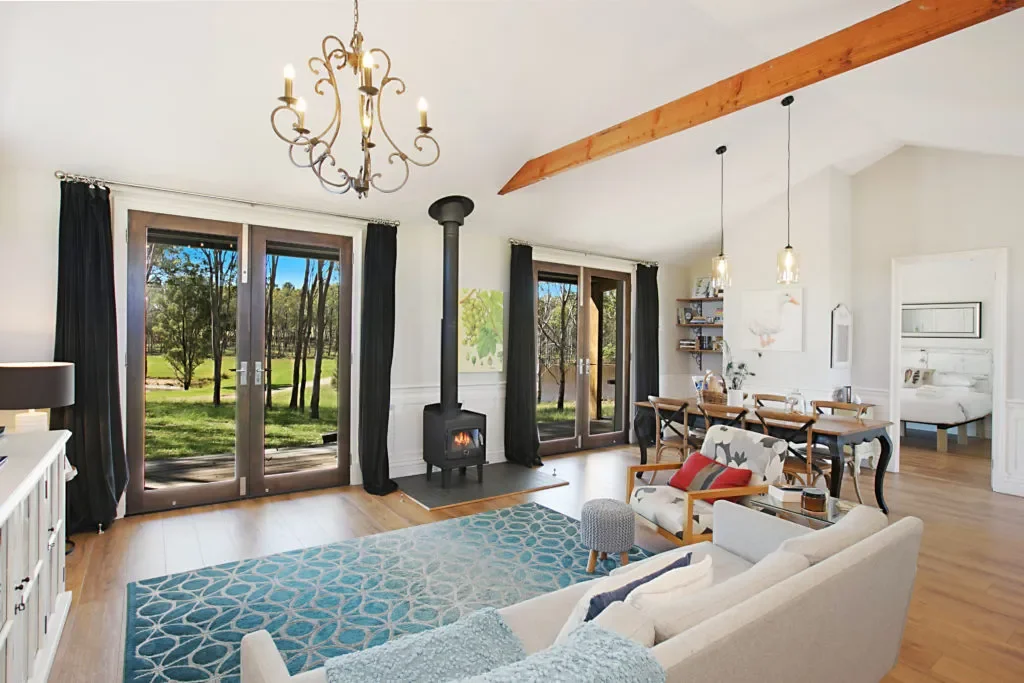 Living room with large glass doors opening to a green outdoor area, a wood-burning stove, a chandelier, a dining table with chairs, and a cozy seating area with a sofa and armchair.