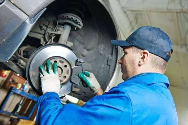 A mechanic in a blue uniform and cap repairing a car's brake system in an auto shop.