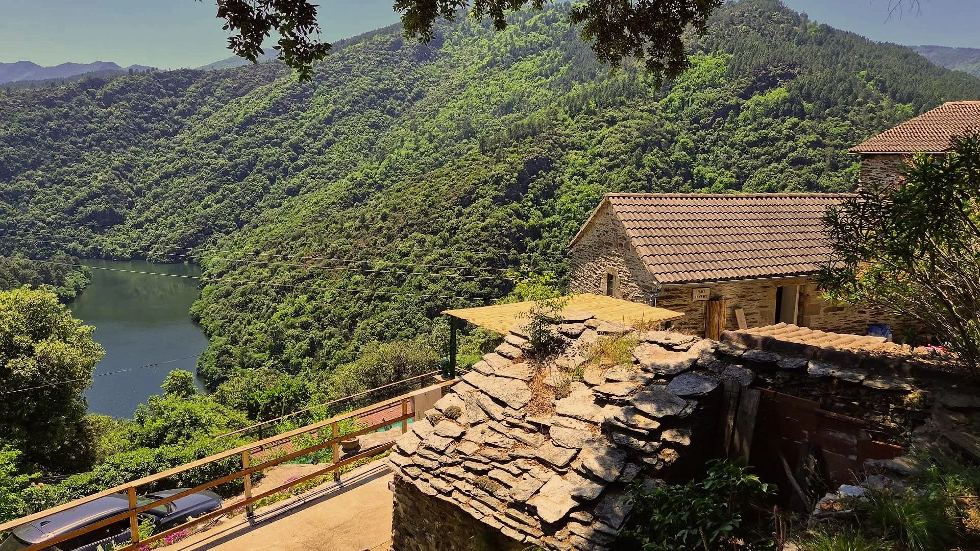 Een huis met een stenen muur en een rode tegelonderkant, gelegen op een heuvel met een uitzicht op een rivier en dichtbegroeide groene bergen.