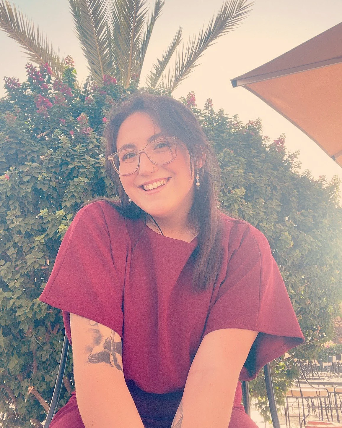 The founder, Lydia, with long dark hair, glasses, and earrings smiling while sitting outdoors at a cafe table, with plants, pink flowers, and an umbrella in the background.