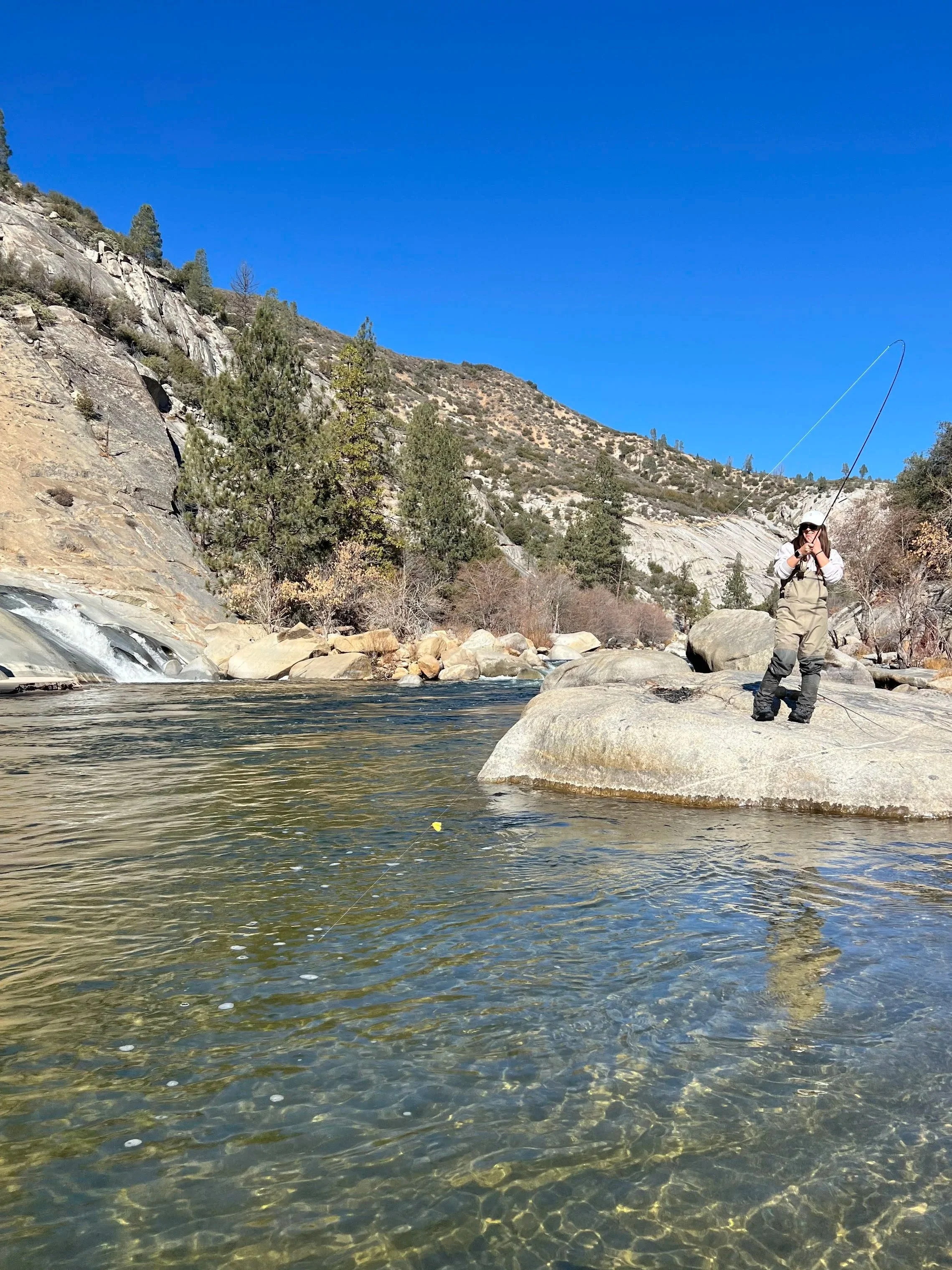 A person dressed in fishing gear standing on rocks by a clear flowing river, holding a fishing rod in a mountainous area with trees and blue sky.