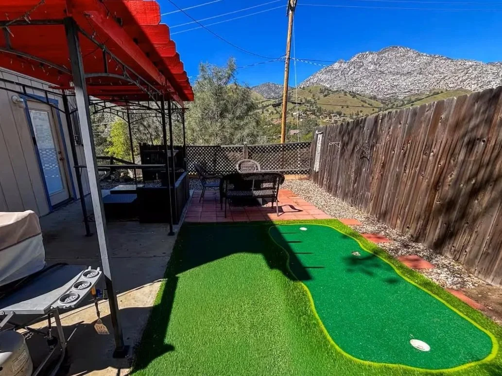 Backyard with artificial grass putting green, outdoor furniture, and a mountain landscape in the background under a clear blue sky.