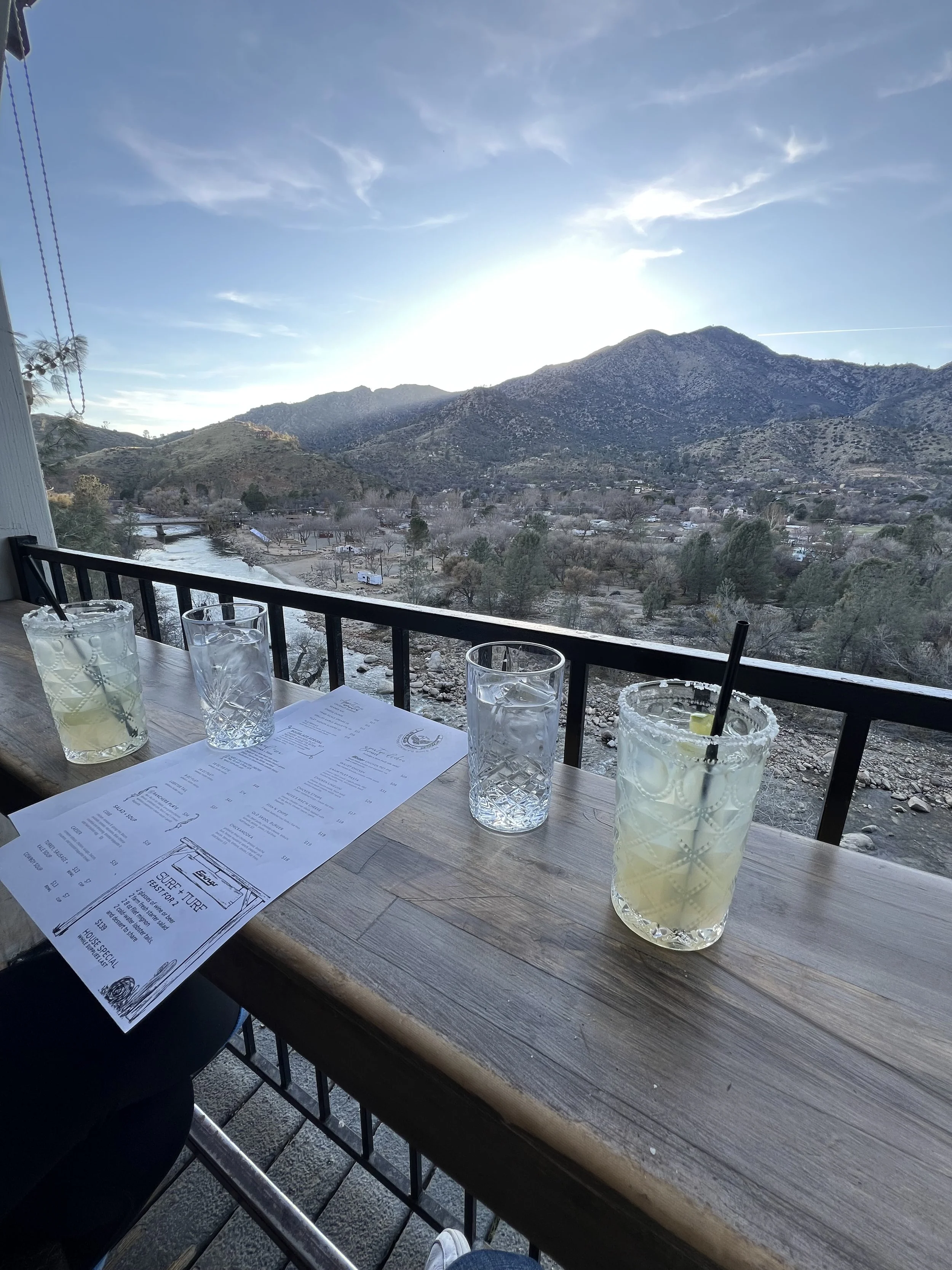 View of a mountain landscape with clear sky, river, and outdoor seating with drinks and a menu on a wooden table.