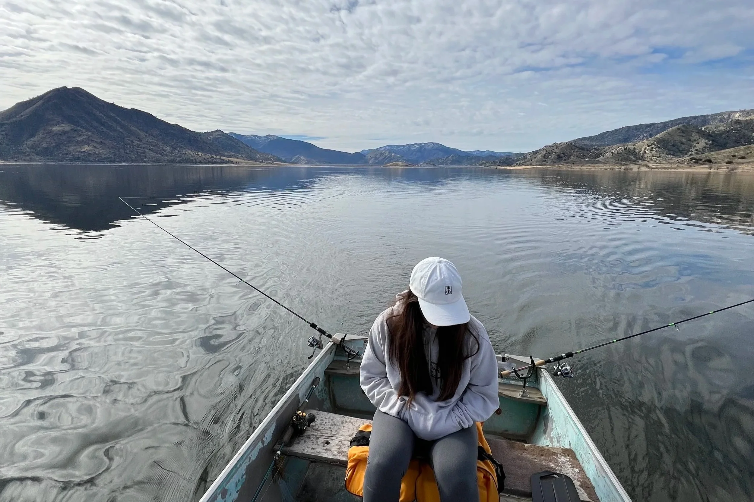 Person wearing a white hat and jacket sitting in a boat on a large, calm lake surrounded by mountains under a cloudy sky, fishing.