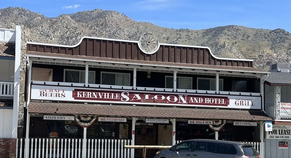 Front view of Kernville Saloon and Hotel, a two-story building with a rustic facade, mountain backdrop, signage for craft beers, whiskey, food, and grill, and a car parked in front.