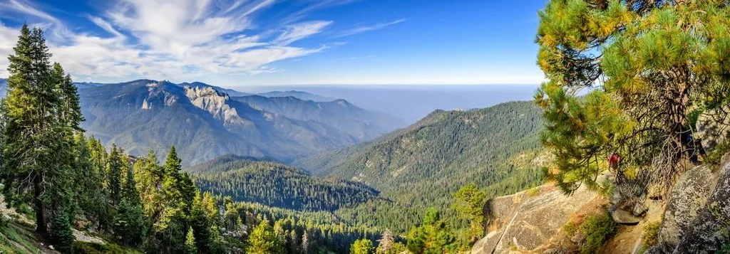 A panoramic view of green forested mountains under a blue sky, with distant mountains and a faint glimpse of the ocean on the horizon.