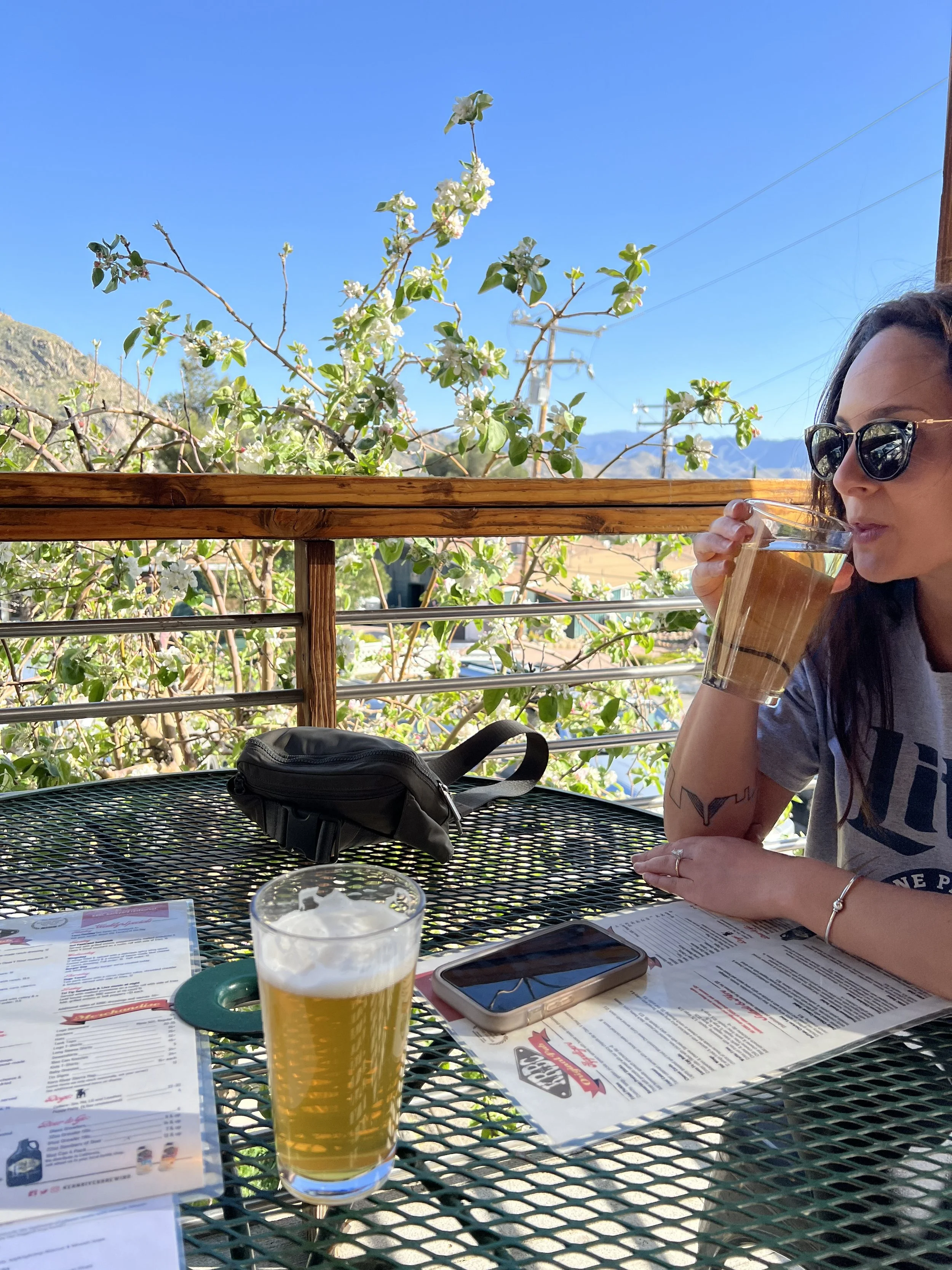 A woman wearing sunglasses and a gray T-shirt is drinking a glass of beer at an outdoor dining area with mountain scenery in the background. The table has menus, a smartphone, and a glass of beer. There are blooming plants and a clear blue sky.