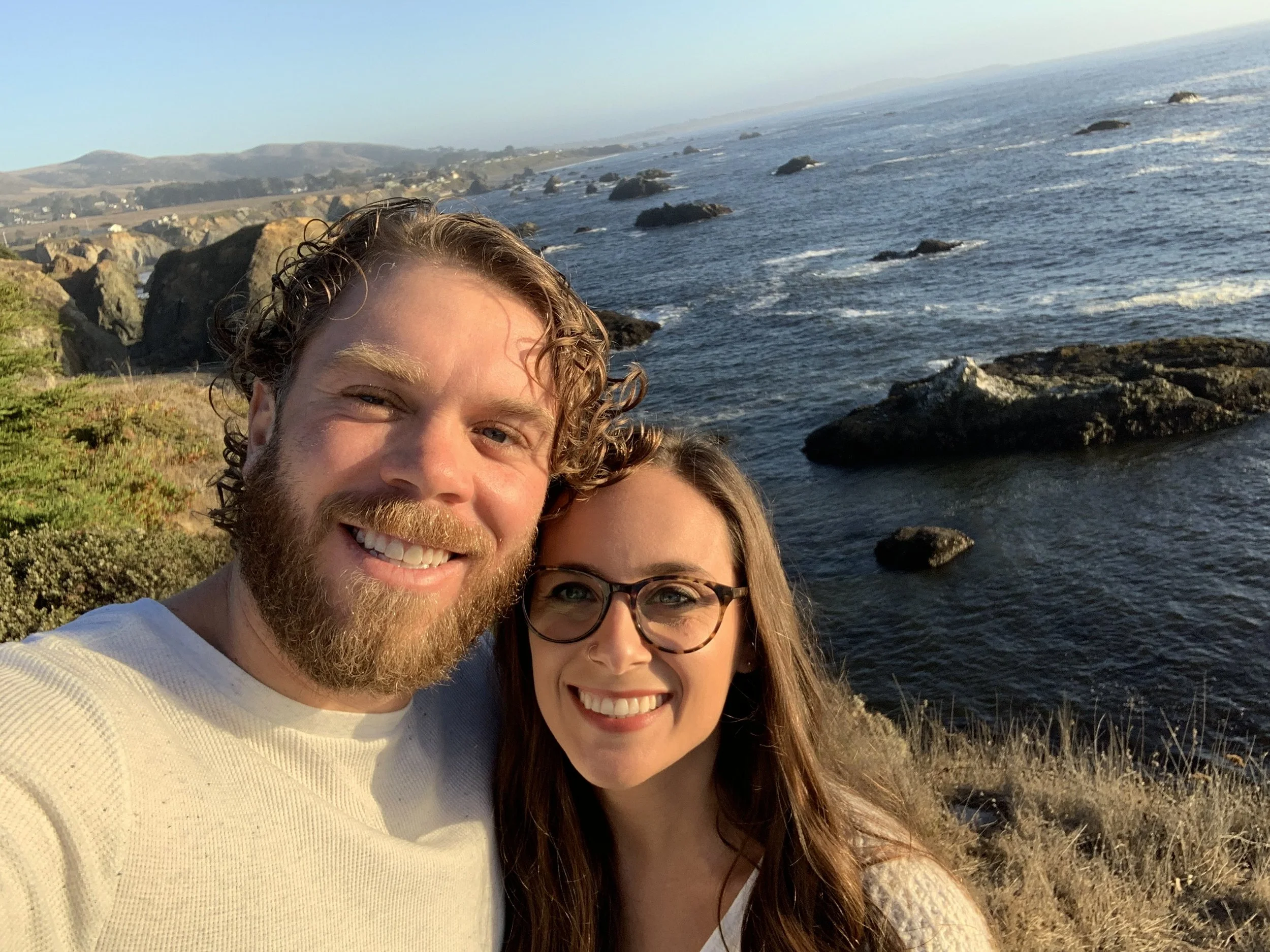 Close-up of a smiling man with curly hair and a beard next to a woman with long hair and glasses, taking a selfie at a rocky coastline during sunset with the ocean in the background.