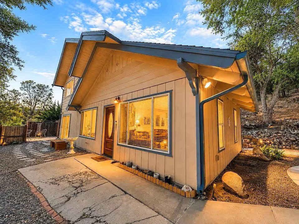 A cozy house with large front windows glowing with warm interior lights. The house has beige siding, a dark gray roof with overhangs, and a wooden front door. The yard has a cracked concrete walkway, a small flower bed, and surrounded by trees and a 