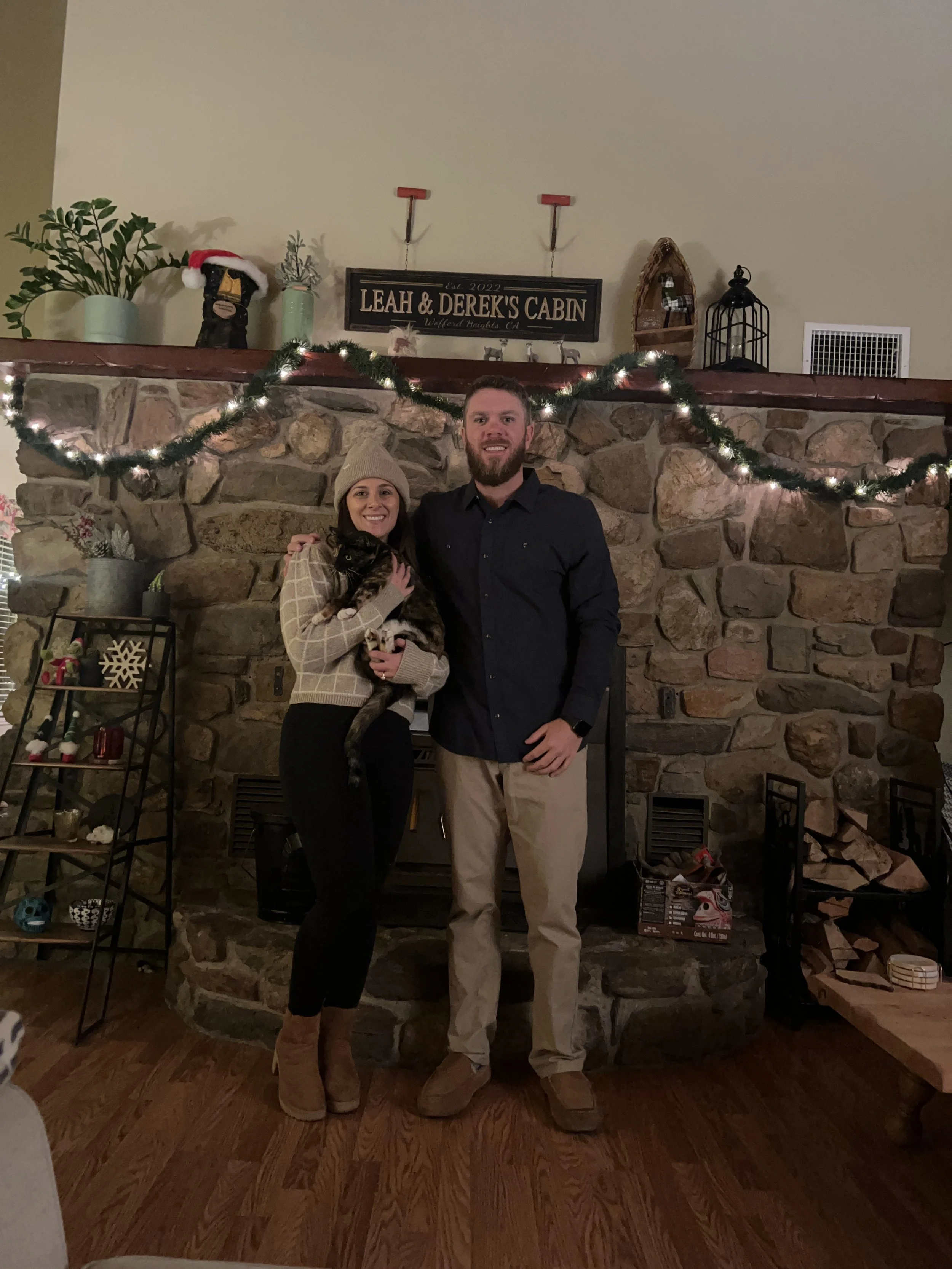 A woman and a man are standing in front of a stone fireplace, smiling. The woman is holding a black cat. The woman is wearing a beige beanie, beige sweater, black pants, and beige shoes. The man is wearing a black shirt and beige pants. The fireplace mantel is decorated with holiday decorations including a string of lights, plants, a holiday-themed hat on a stuffed animal, and various other ornaments. There is a sign on the wall that reads 'Leah & Derek's Cabin, Wofford Heights CA.'