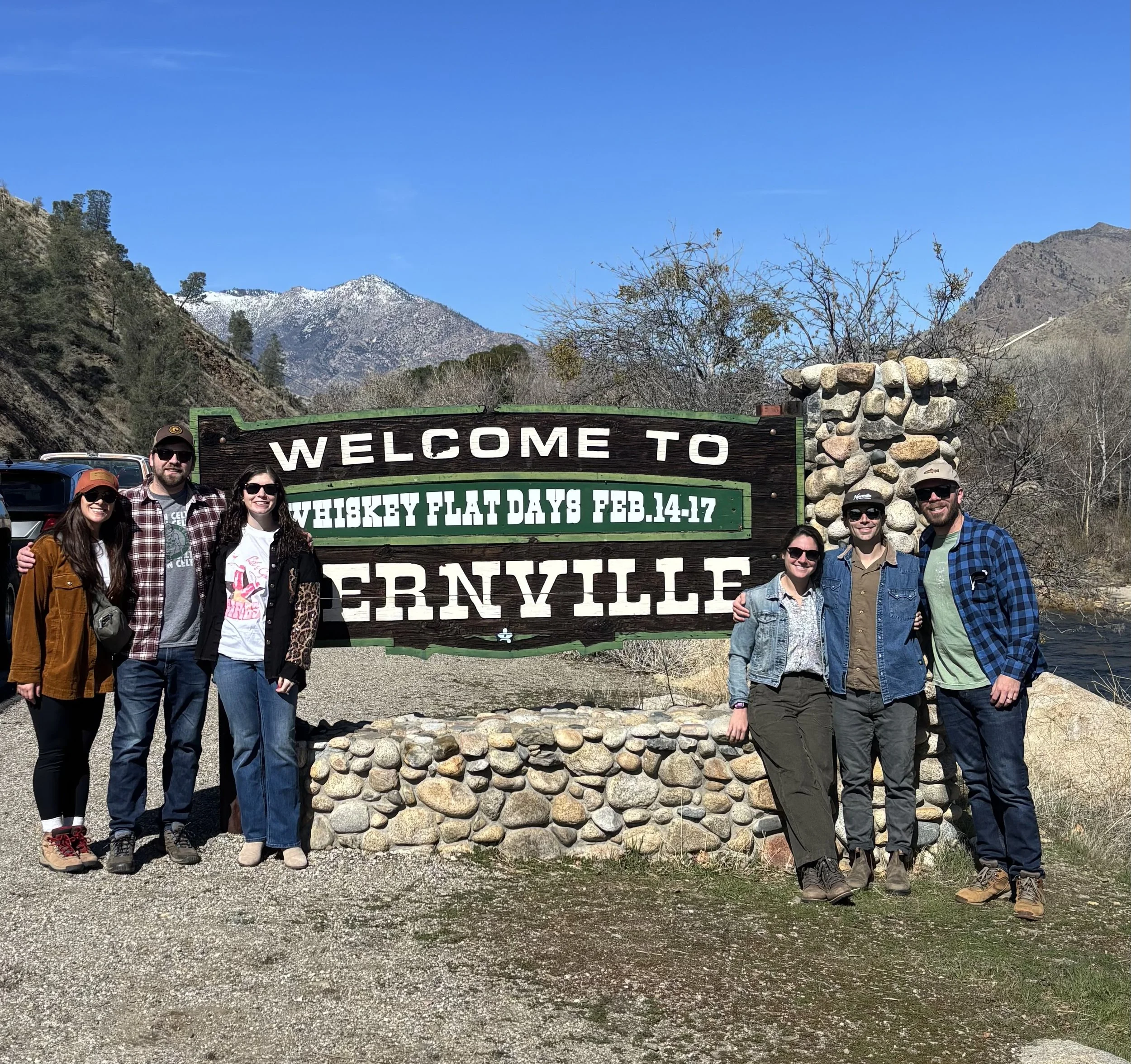 A group of eight people standing in front of a mountain landscape and a wooden sign that reads 'Welcome to Whiskey Flat Days Feb. 14-17 Kernville.' They are wearing casual outdoor clothing and sunglasses, and appear to be smiling and enjoying a sunny day.