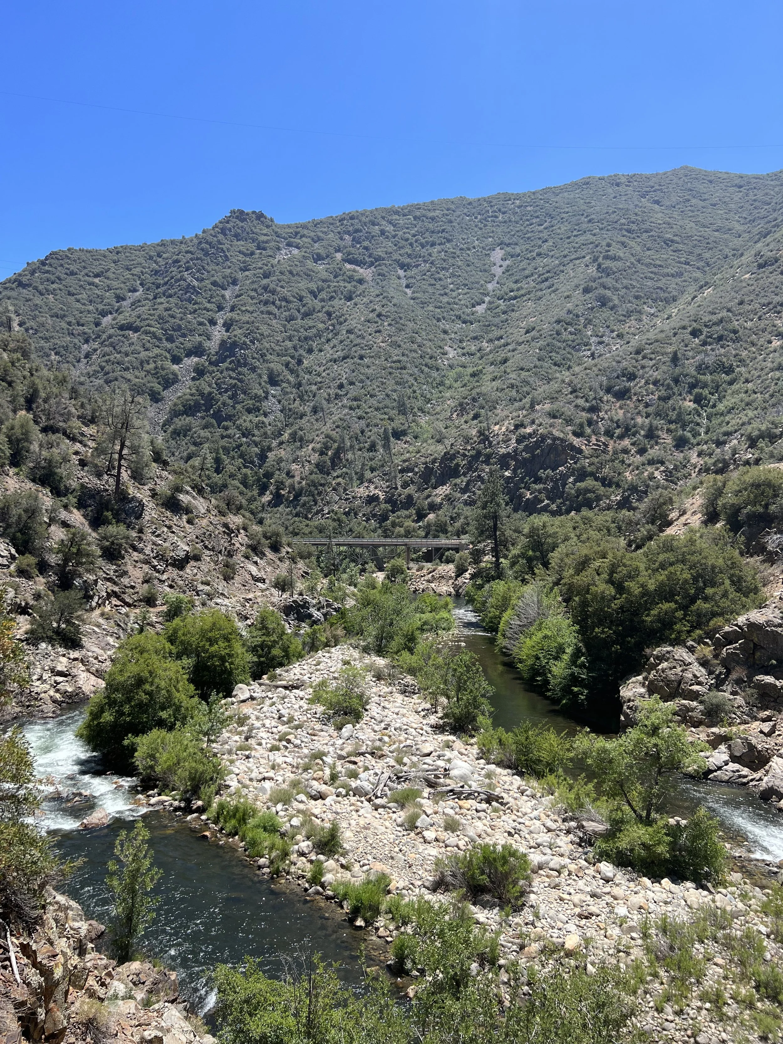 A scenic mountain landscape with a river flowing through it, surrounded by lush green trees and rocky terrain under a clear blue sky, with a bridge crossing the river in the distance.