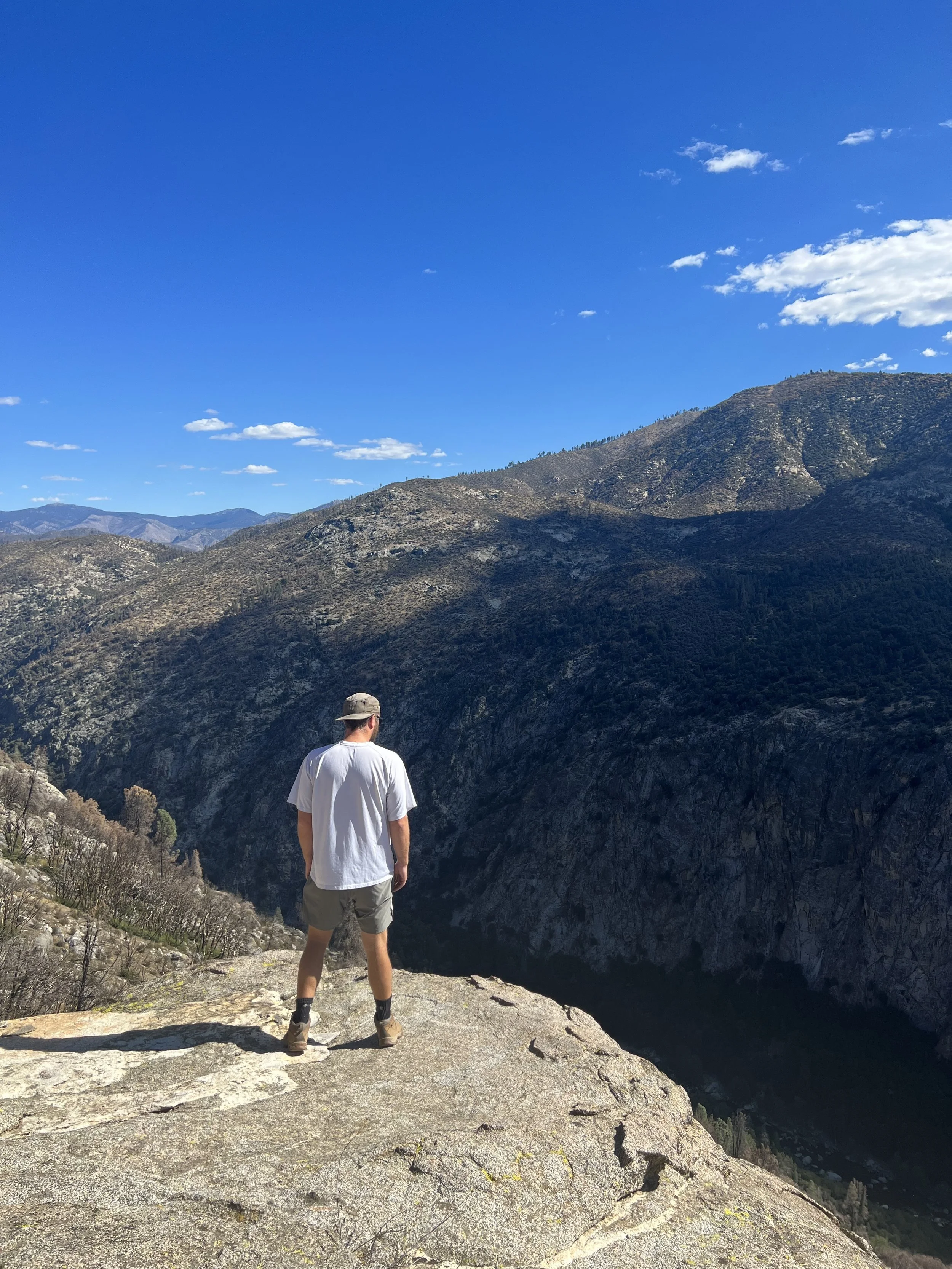 A man stands on a large rock overlooking a deep canyon with rugged, forested mountains under a bright blue sky with scattered clouds.
