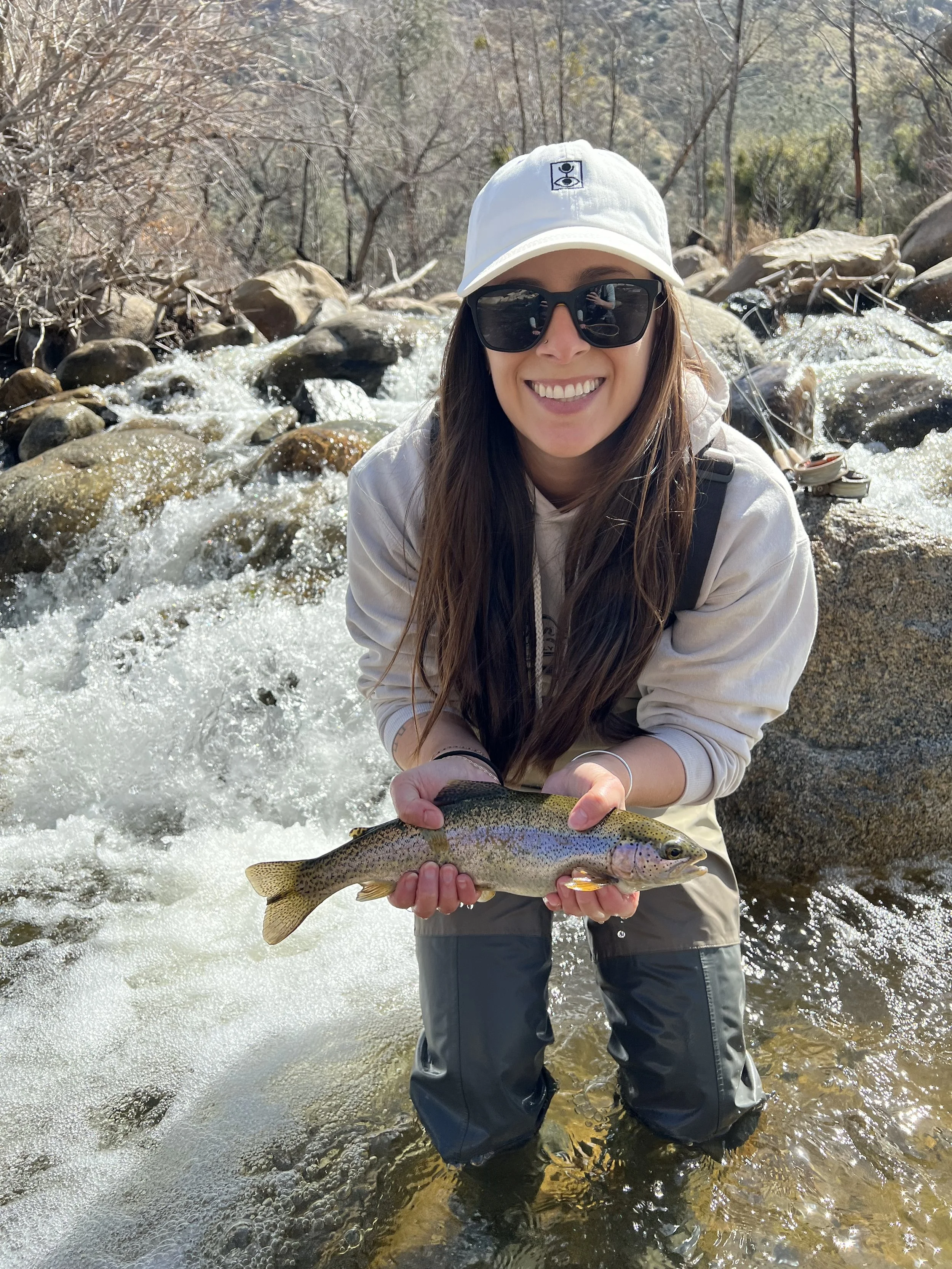 A woman outdoors in a river holding a rainbow trout fish she caught, wearing sunglasses, a white hat, a light hoodie, and waders, smiling with a natural landscape of rocks, water, and trees in the background.