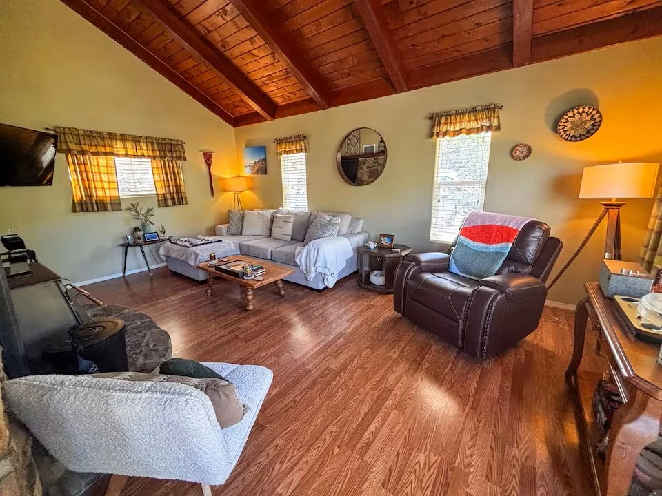Living room with wood ceiling, yellow walls, hardwood floor, and several windows with plaid valances. Contains a beige sectional sofa, a black leather recliner with a multicolored throw, a white armchair, and various side tables and lamps.