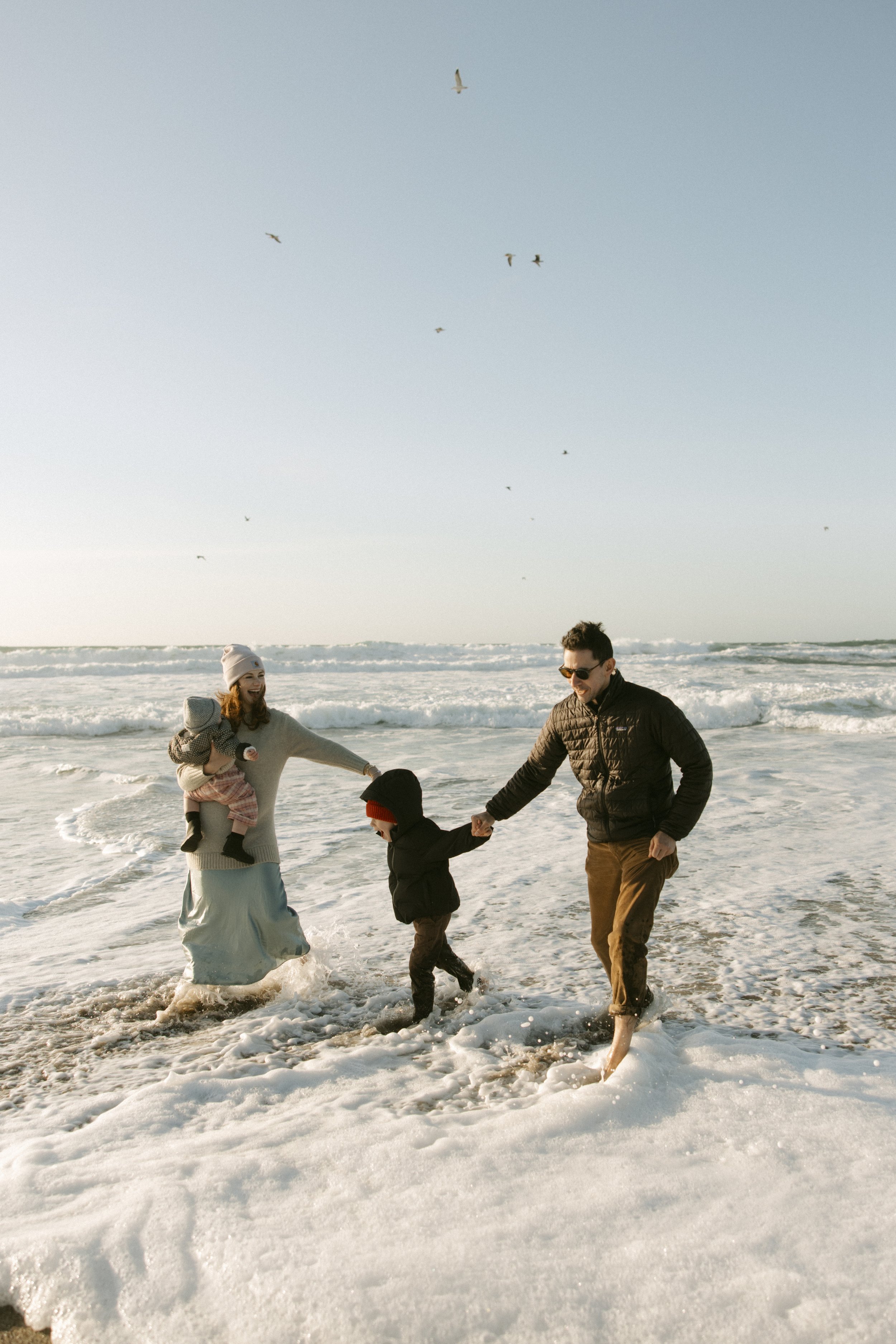 A family of four playing in the surf on a beach with seagulls flying overhead and a clear sky.