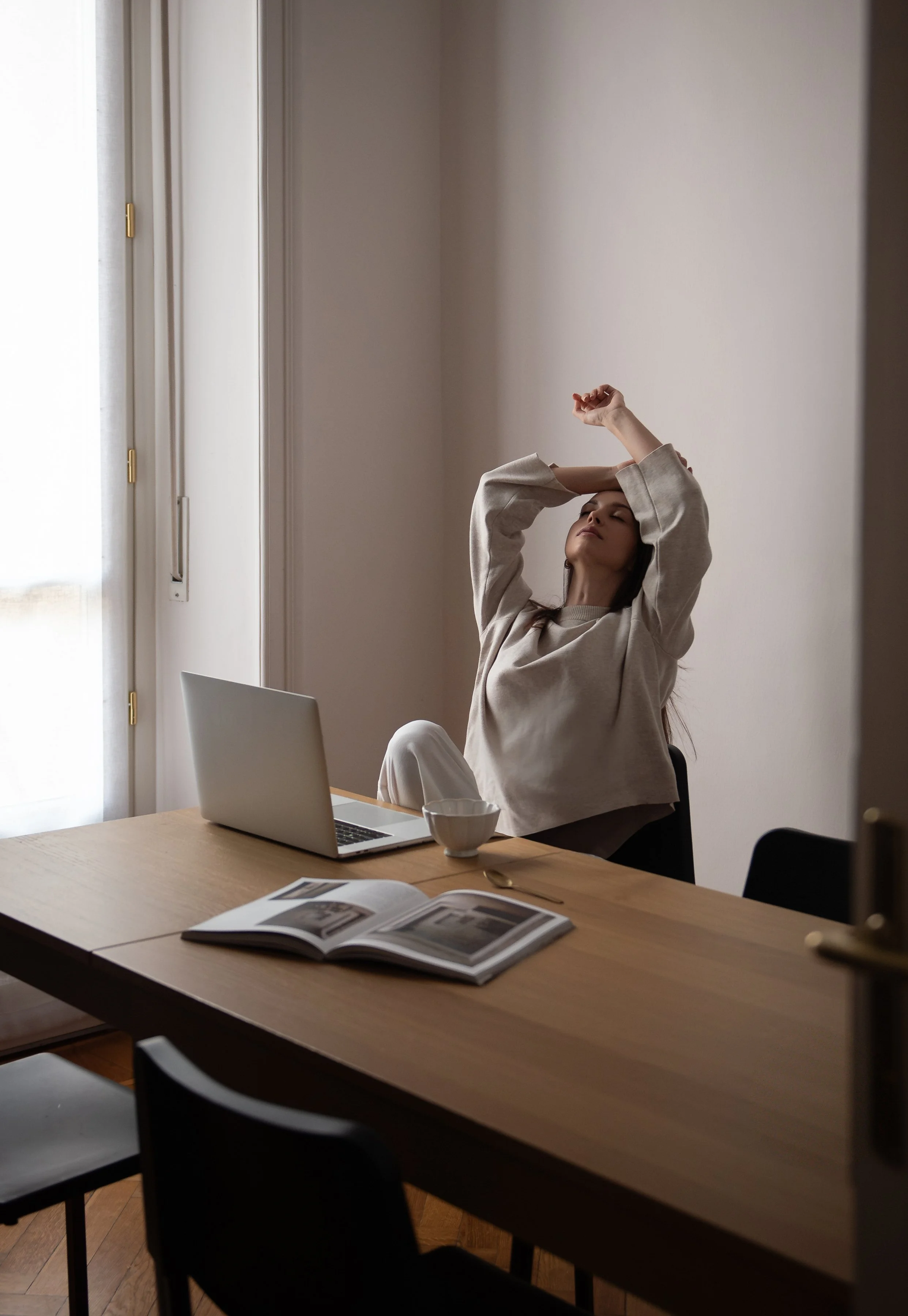 A woman with long dark hair sitting at a wooden table, stretching with her eyes closed and hands behind her head in a well-lit room with a white wall and a window. There is a laptop, an open magazine, and a cup on the table.