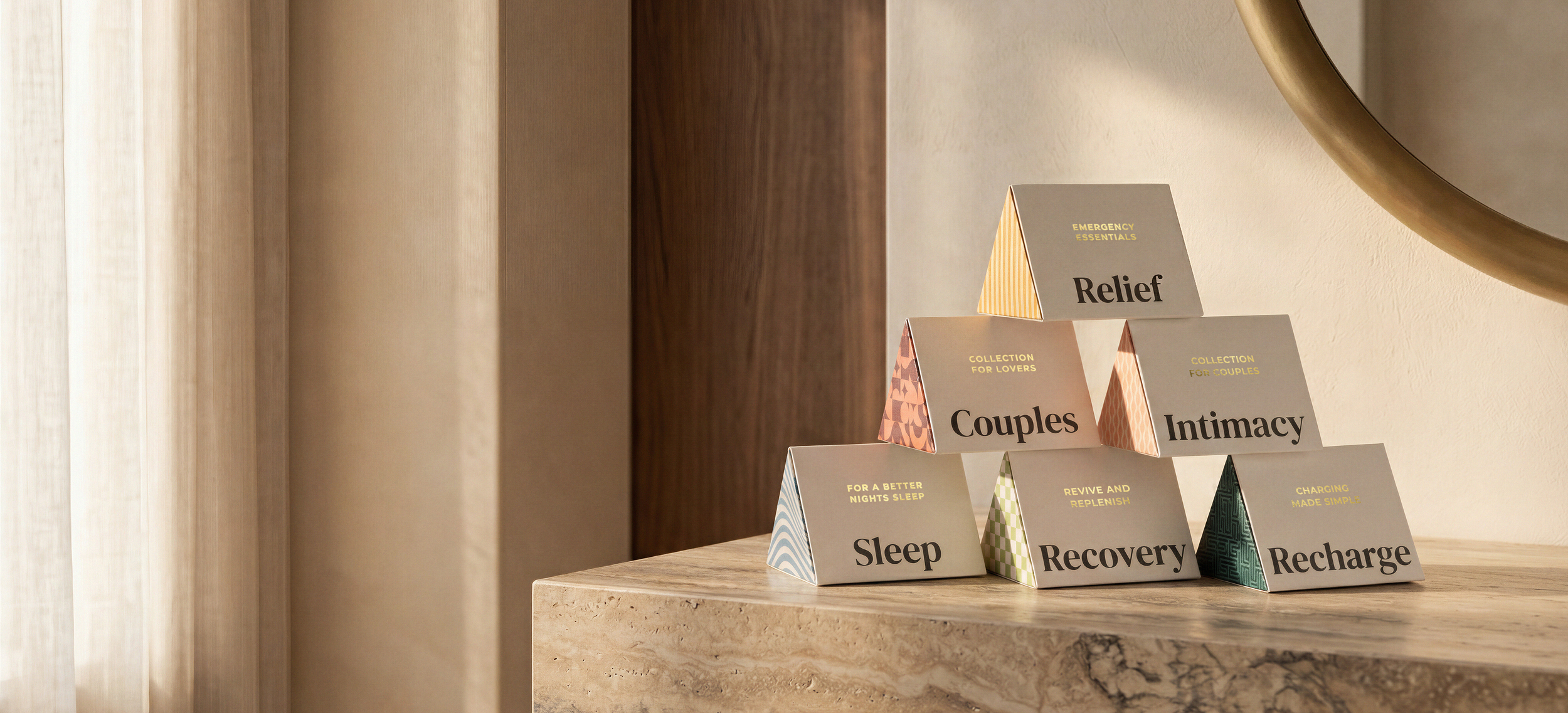 A pyramid arrangement of boxes with wellness and self-care words, including 'Sleep,' 'Recovery,' 'Recharge,' 'Couples,' 'Intimacy,' 'Relief,' and 'Emergency Essentials,' on a wooden surface next to a wall and a mirror.