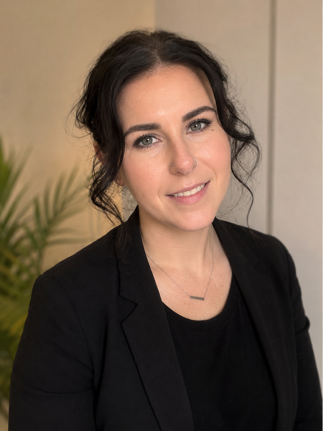 A woman with dark, wavy hair, light skin, green eyes, wearing a black blazer and a silver bar necklace, smiling in front of a neutral background with a plant partially visible to her left.