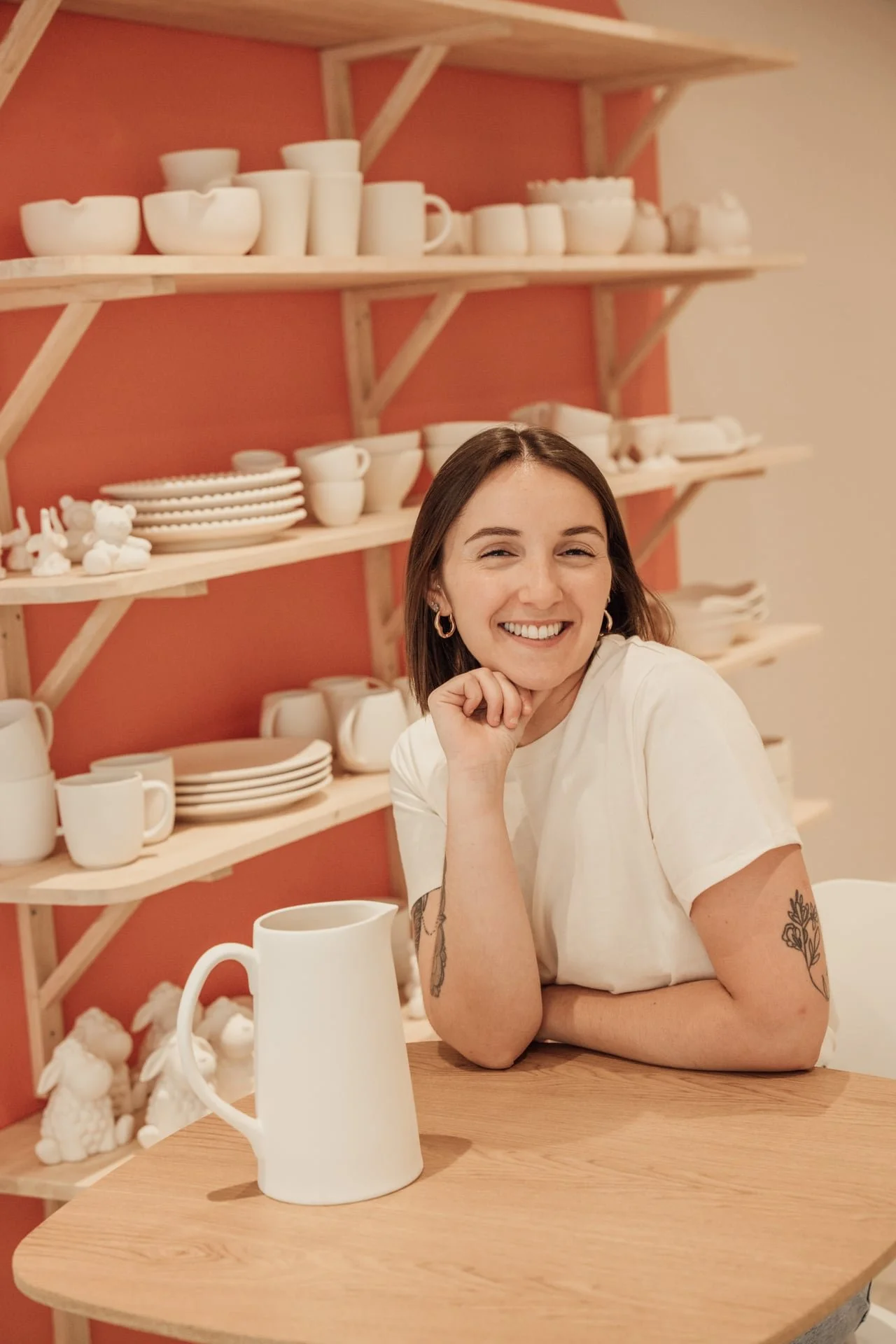 Une jeune femme souriante avec des tatouages sur les bras, assise à une table en bois dans une cuisine ou un espace de vaisselle avec des étagères en bois et de la vaisselle blanche.