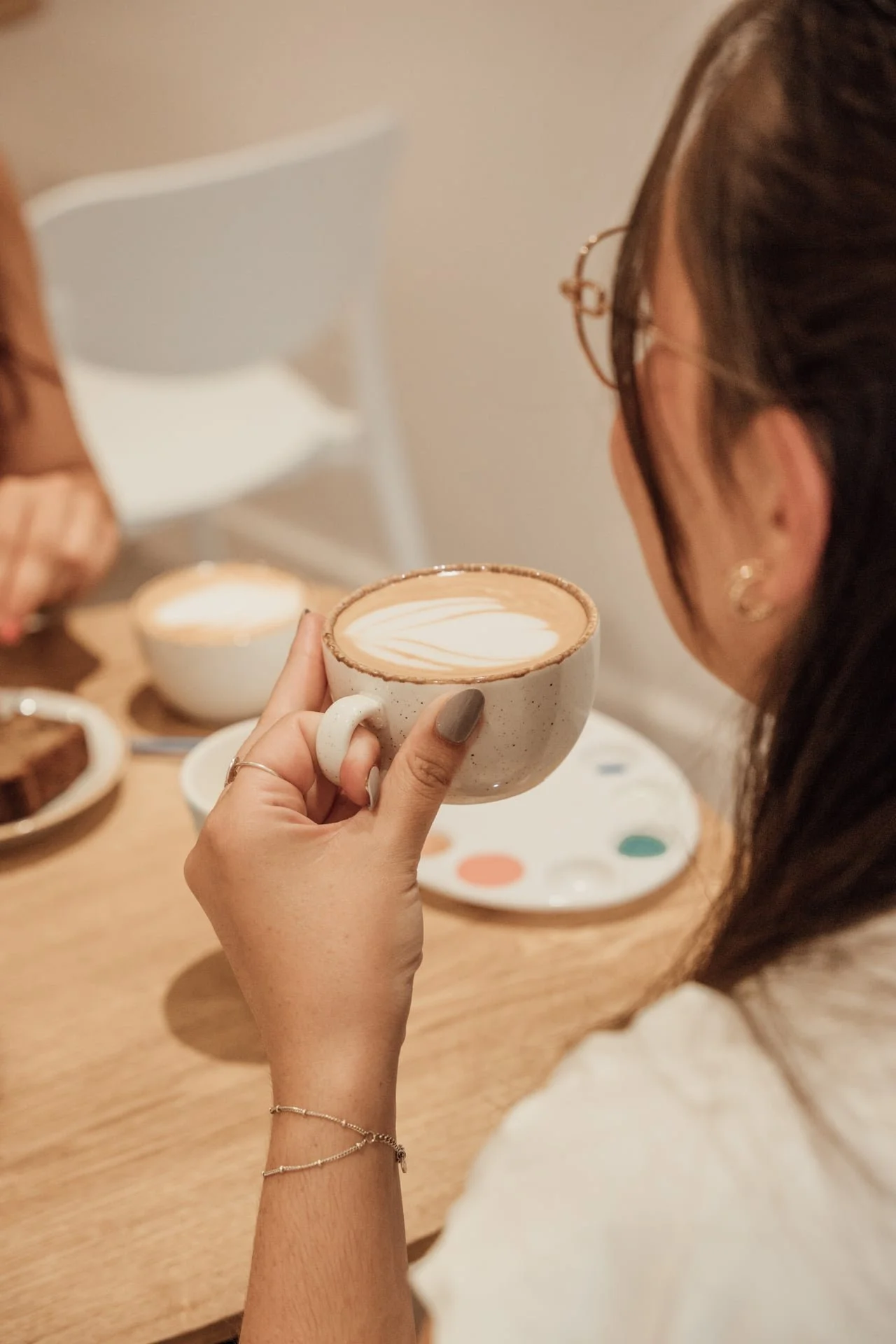 Une personne tient une tasse de café ou de cappuccino avec du latte art, assise à une table en bois, avec des plats et de la vaisselle en arrière-plan.