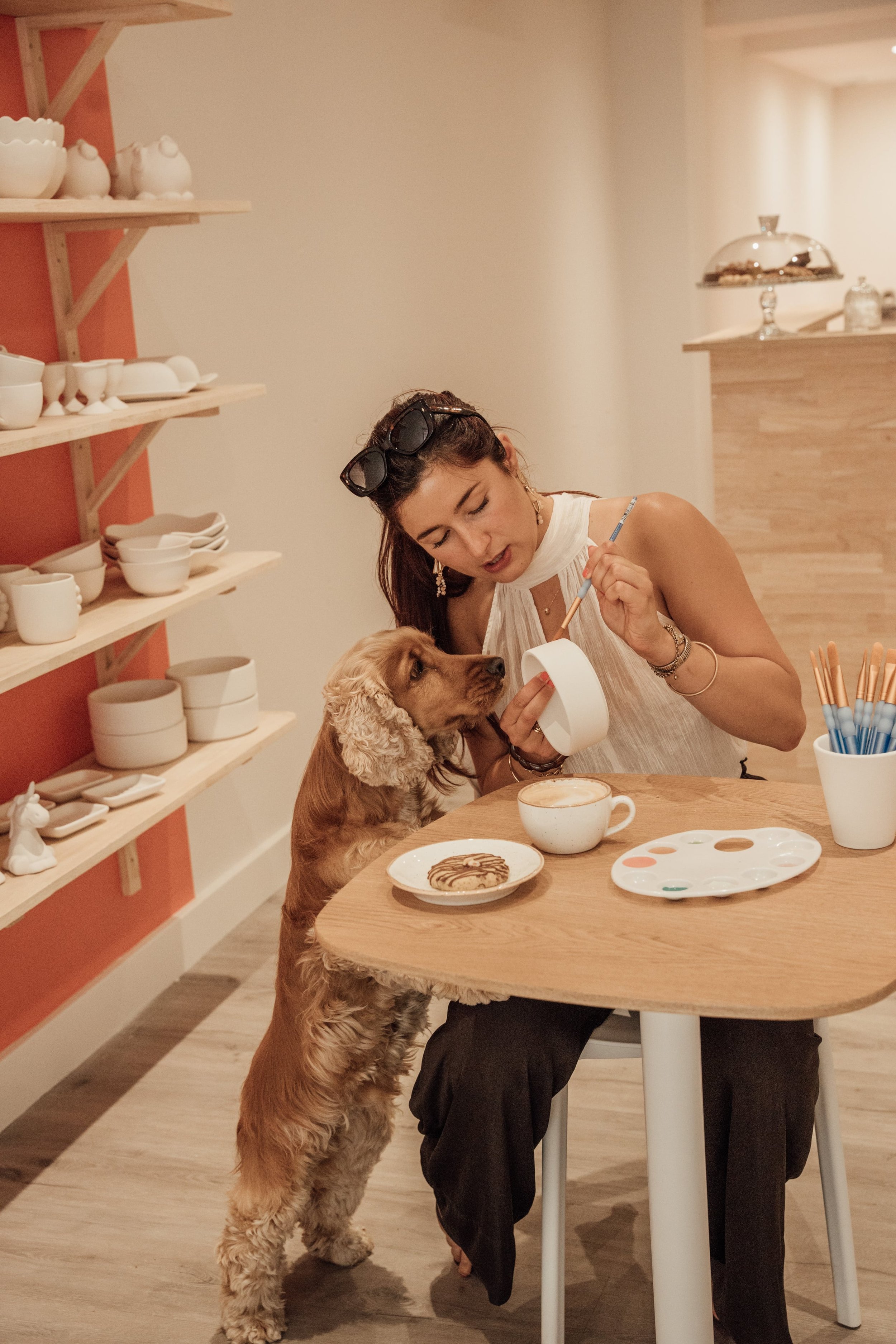 Une femme avec un chien est assise à une table dans un café. La femme tient un pinceau et un mug, probablement en train de peindre ou décorer un objet blanc. La scène est chaleureuse avec des étagères remplies de vaisselle en arrière-plan.
