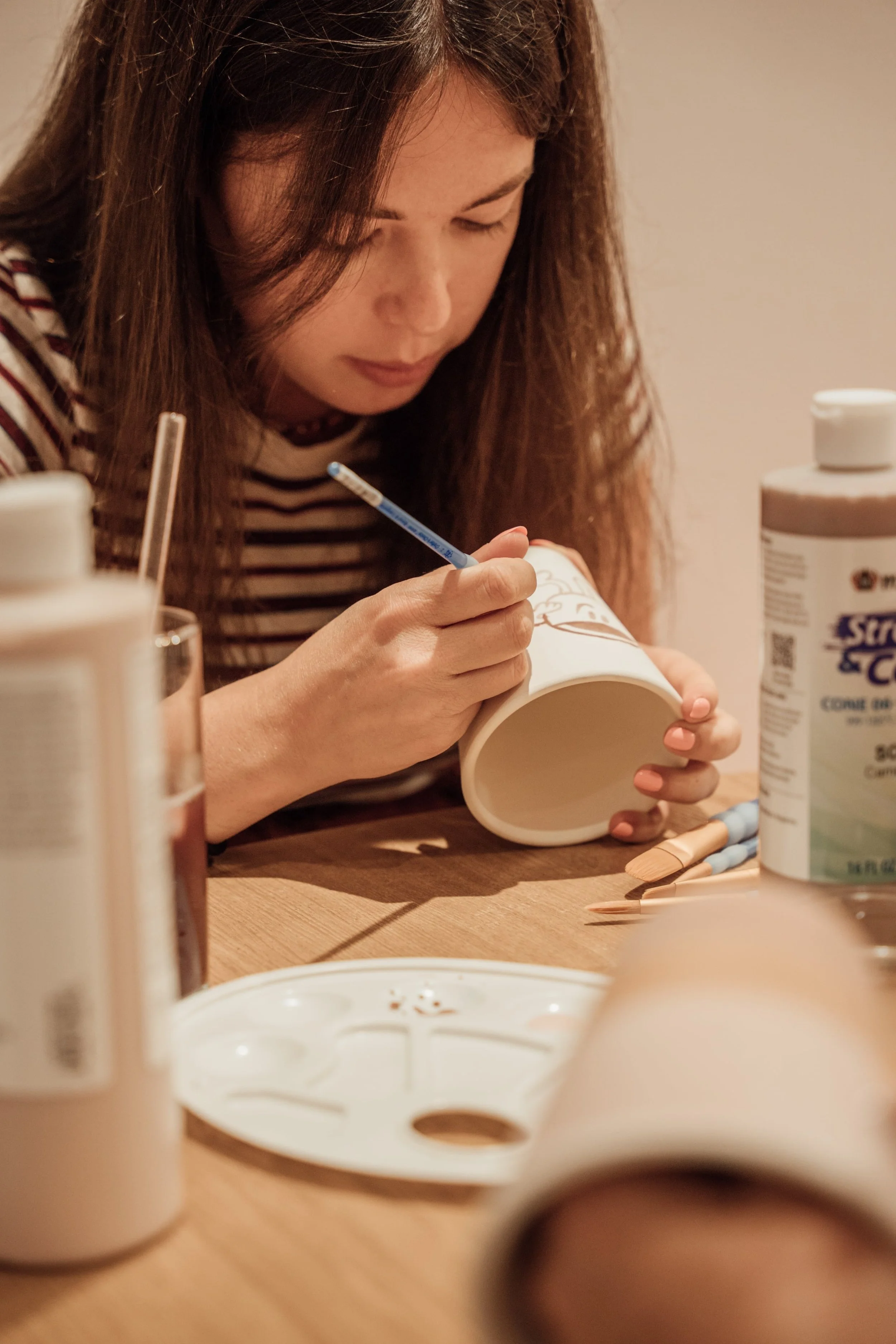 Une femme peignant un récipient en poterie avec des crayons de couleur, entourée de bouteilles de peinture sur une table.