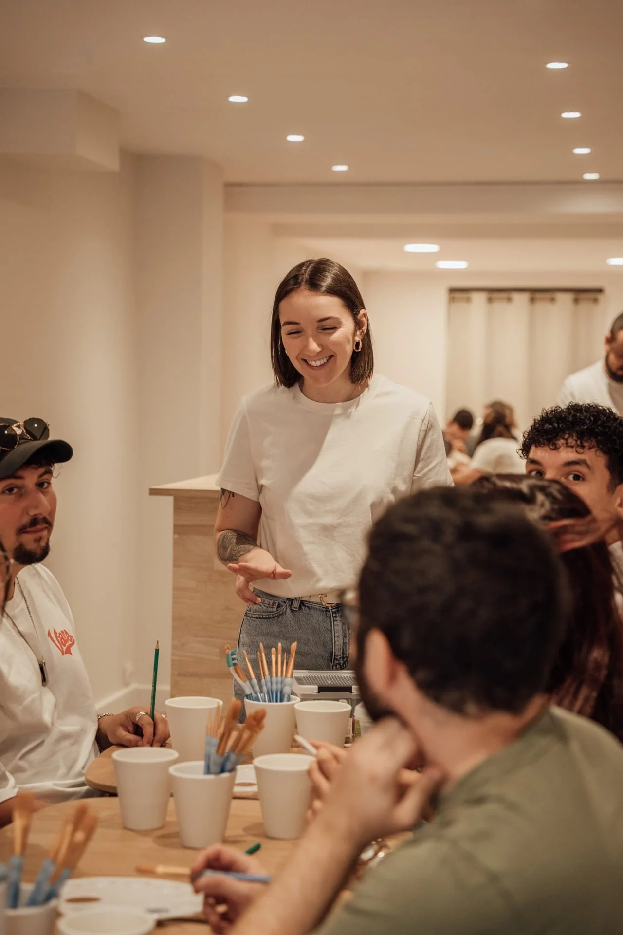 Groupe de personnes autour d'une table, une femme souriante en t-shirt blanc se tenant debout, en arrière-plan, dans une pièce éclairée avec plafond à LED.