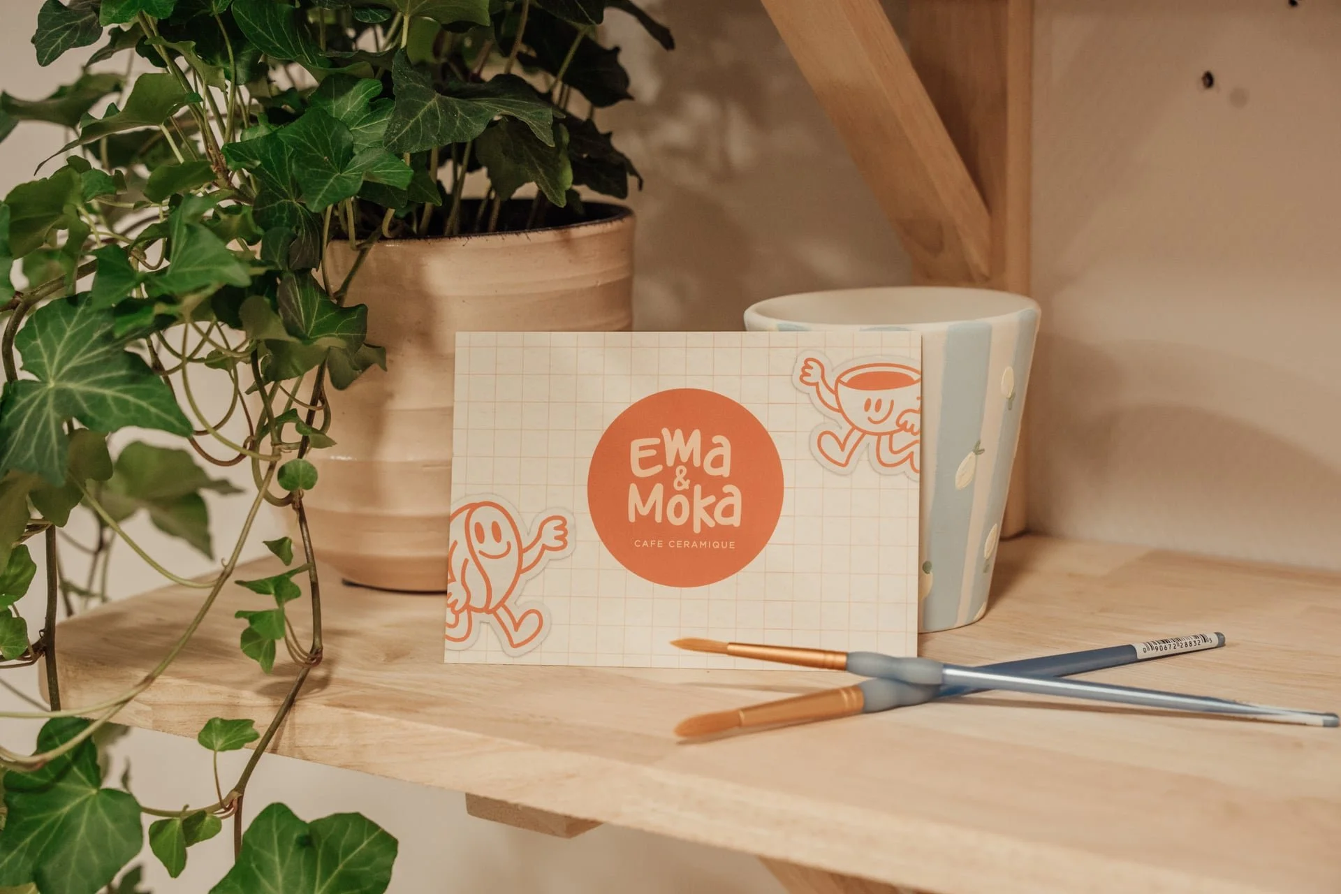 Une plante verte en pot en céramique beige sur une étagère en bois, à côté d'une tasse en céramique avec un dessin de personnage sympathique, une carte avec le texte 'Ewa & Moka Cafe Ceramique' et deux stylos sur une table en bois.