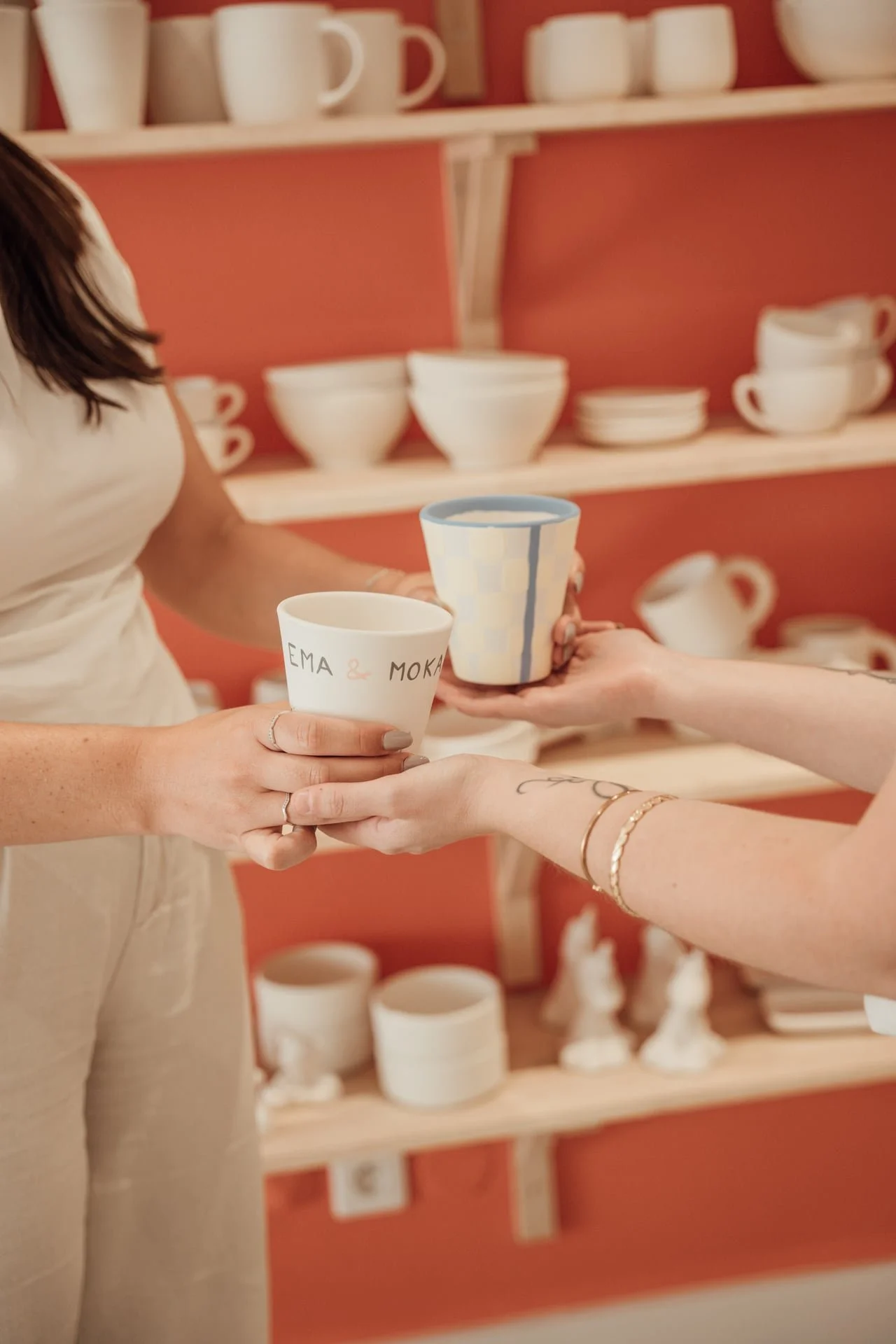 Deux personnes échangent des tasses en céramique, dans une boutique de vaisselle, avec des étagères de tasses et de bols en arrière-plan, dans une ambiance chaleureuse.