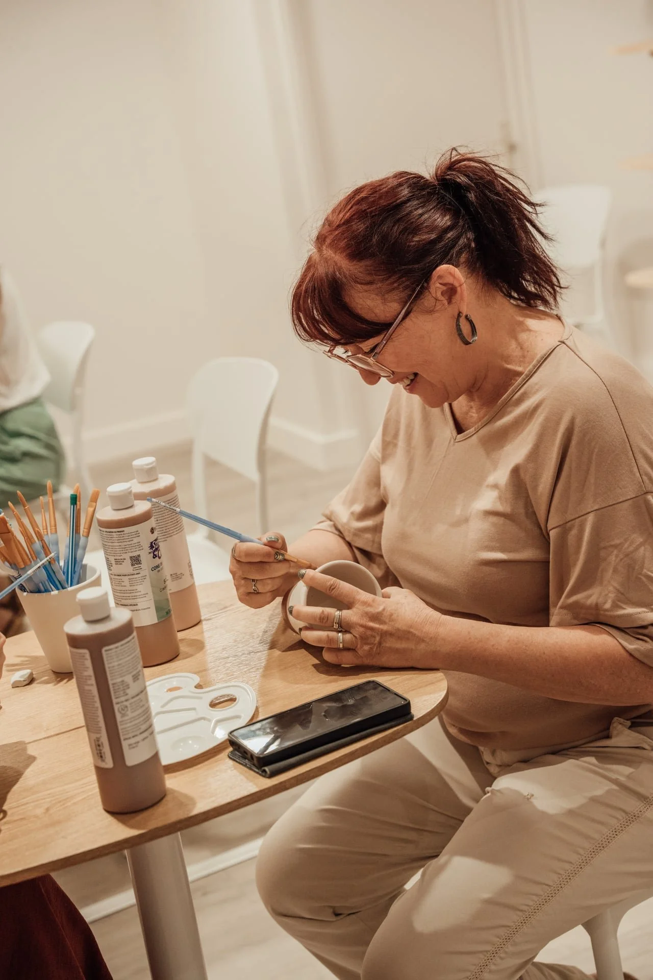 Une femme assise à une table en train de peindre un mug avec des peintures et des pinceaux. Divers tubes de peinture sont sur la table, ainsi qu'un pinceau dans sa main. Elle porte des lunettes, des boucles d'oreilles et un haut beige.