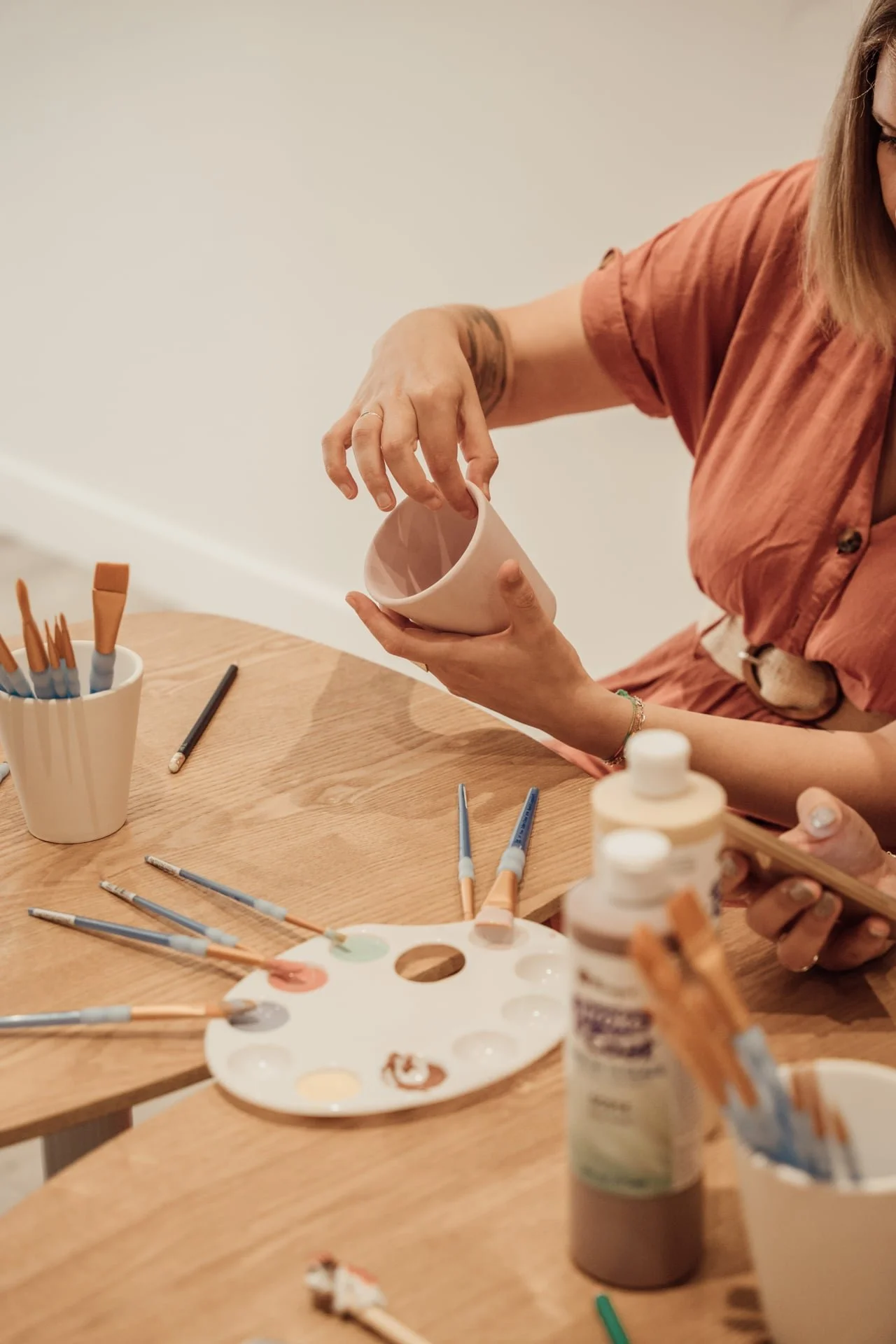 Personne en train de peindre une tasse en céramique avec des pinceaux et des peintures sur une table en bois, entourée de fournitures pour la peinture.