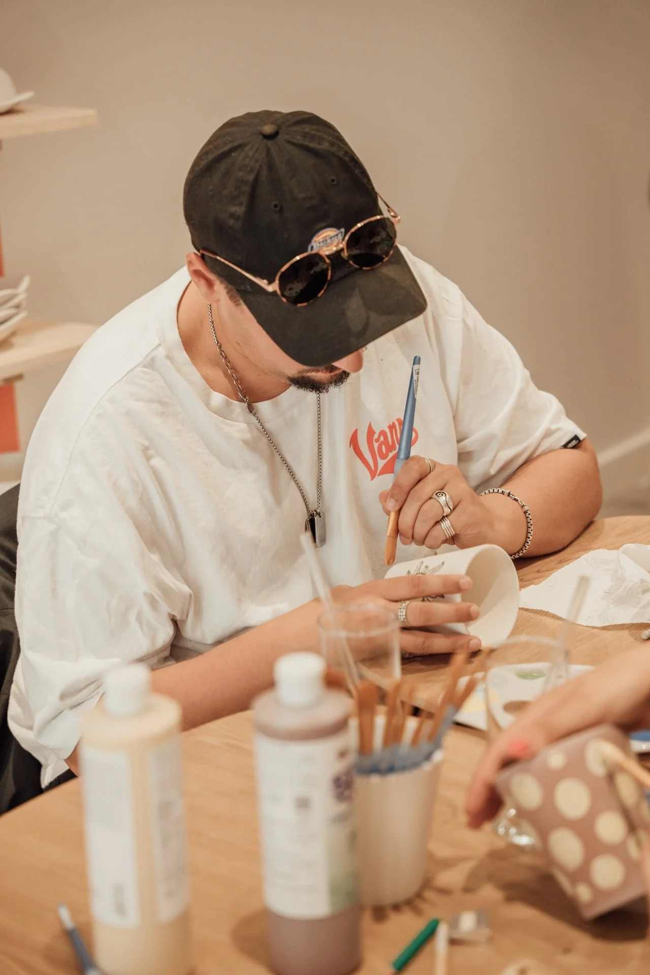 Un jeune homme portant une casquette noire et des lunettes de soleil posées dessus, en train de peindre sur un objet blanc avec un pinceau, assis à une table avec des bouteilles de peinture, des pinceaux et d'autres outils artistiques.
